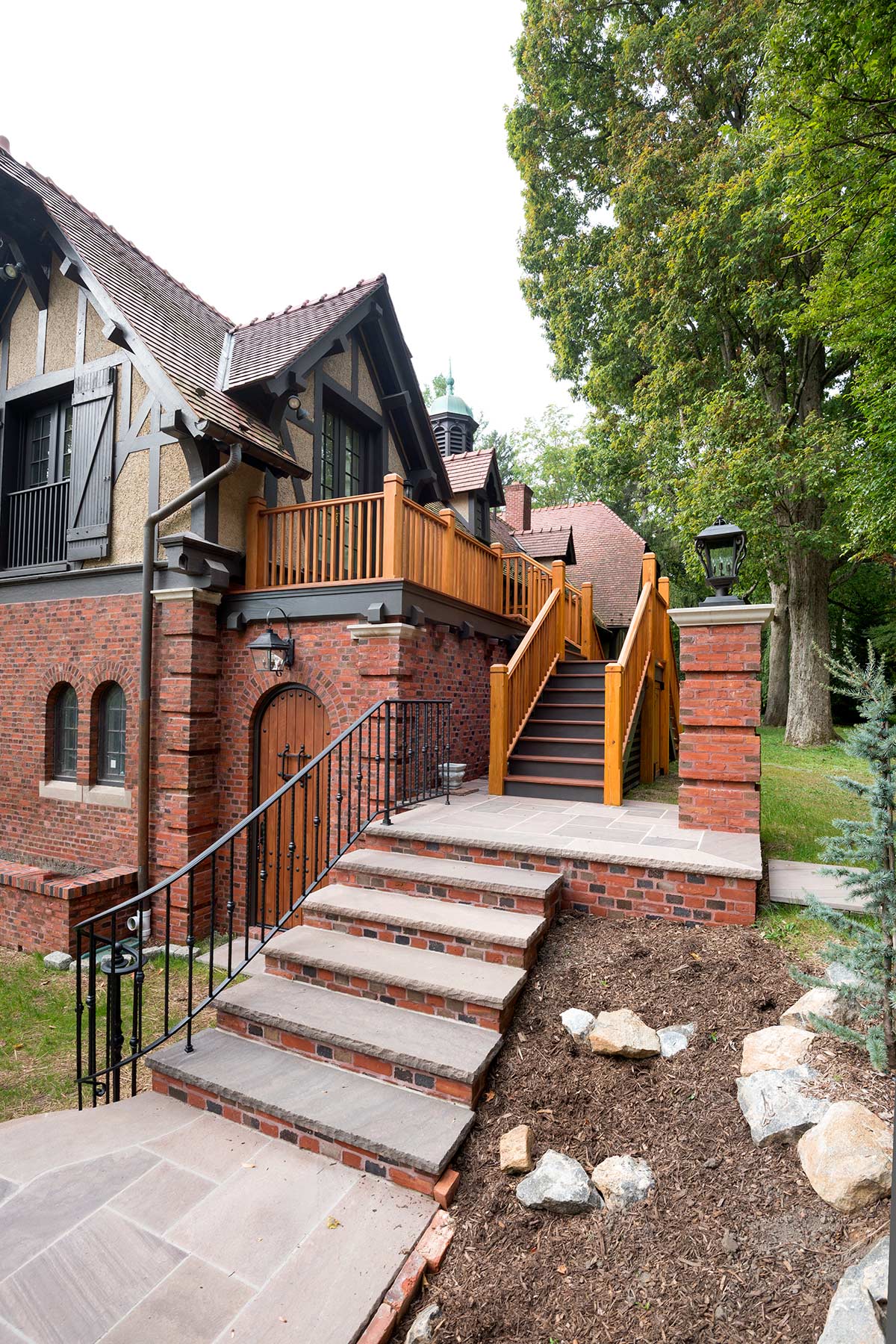 A brick house with a wooden door, balcony, and brick steps leading up, surrounded by trees and landscaping.