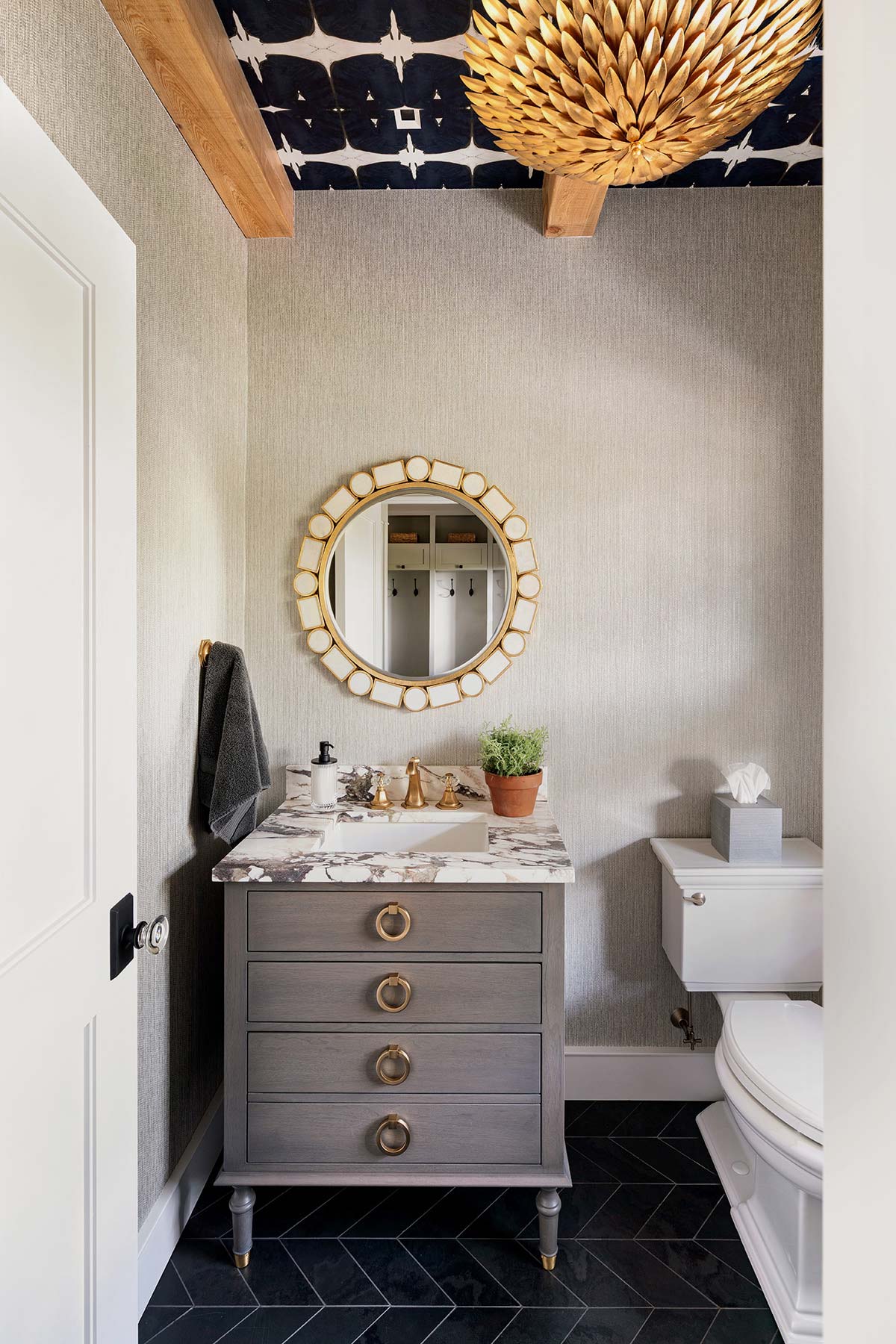 Stylish bathroom with a gray vanity, round gold mirror, black herringbone floor, and a bold patterned ceiling with a gold chandelier.