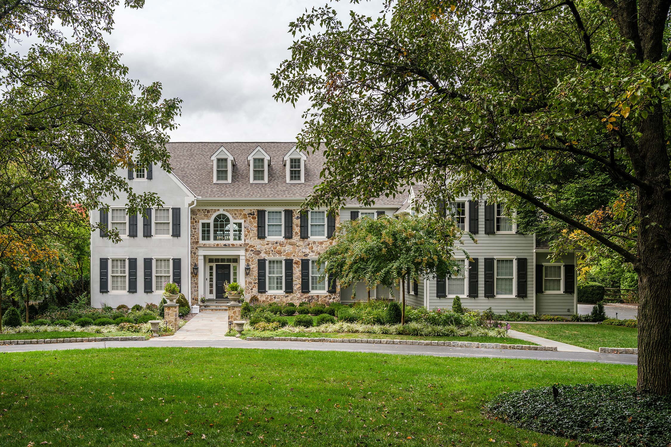 Large two-story house with stone and siding exterior, black shutters, dormer windows, and a well-maintained lawn with trees.