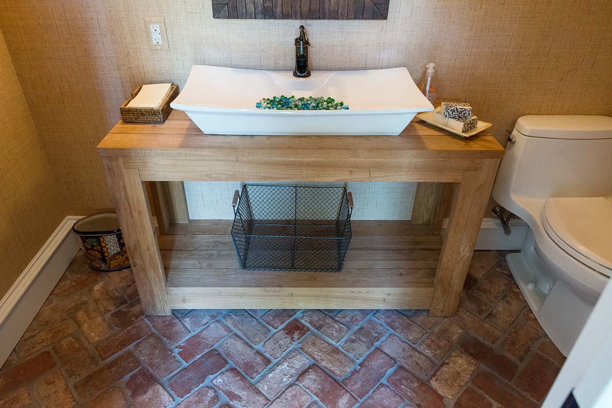 Rustic bathroom with wooden vanity, white sink adorned with colorful stones, brick floor, and modern toilet.