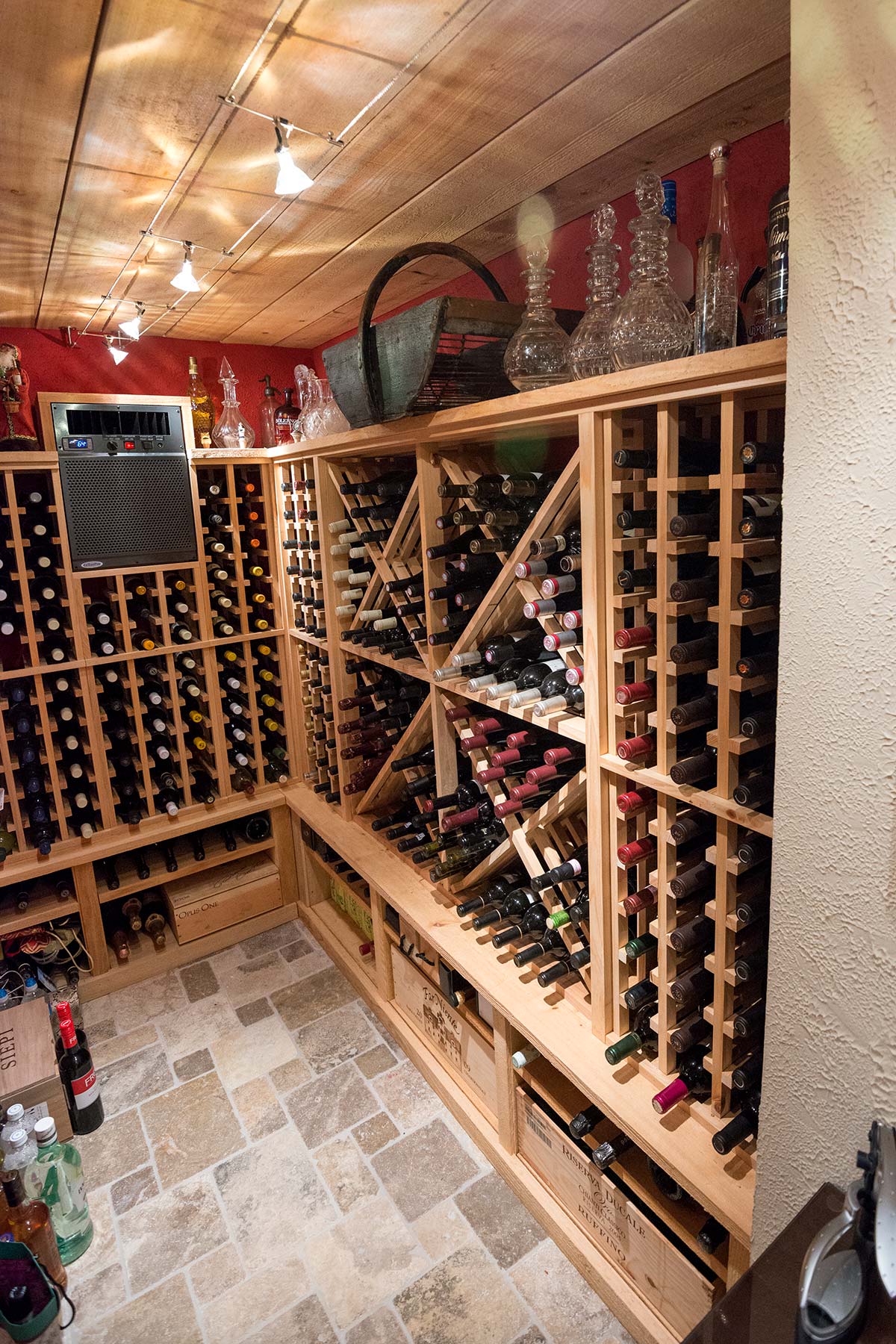 Cozy wine cellar with wooden racks, assorted bottles, and decorative decanters. Features tiled floors, track lighting, and storage crates along the walls.