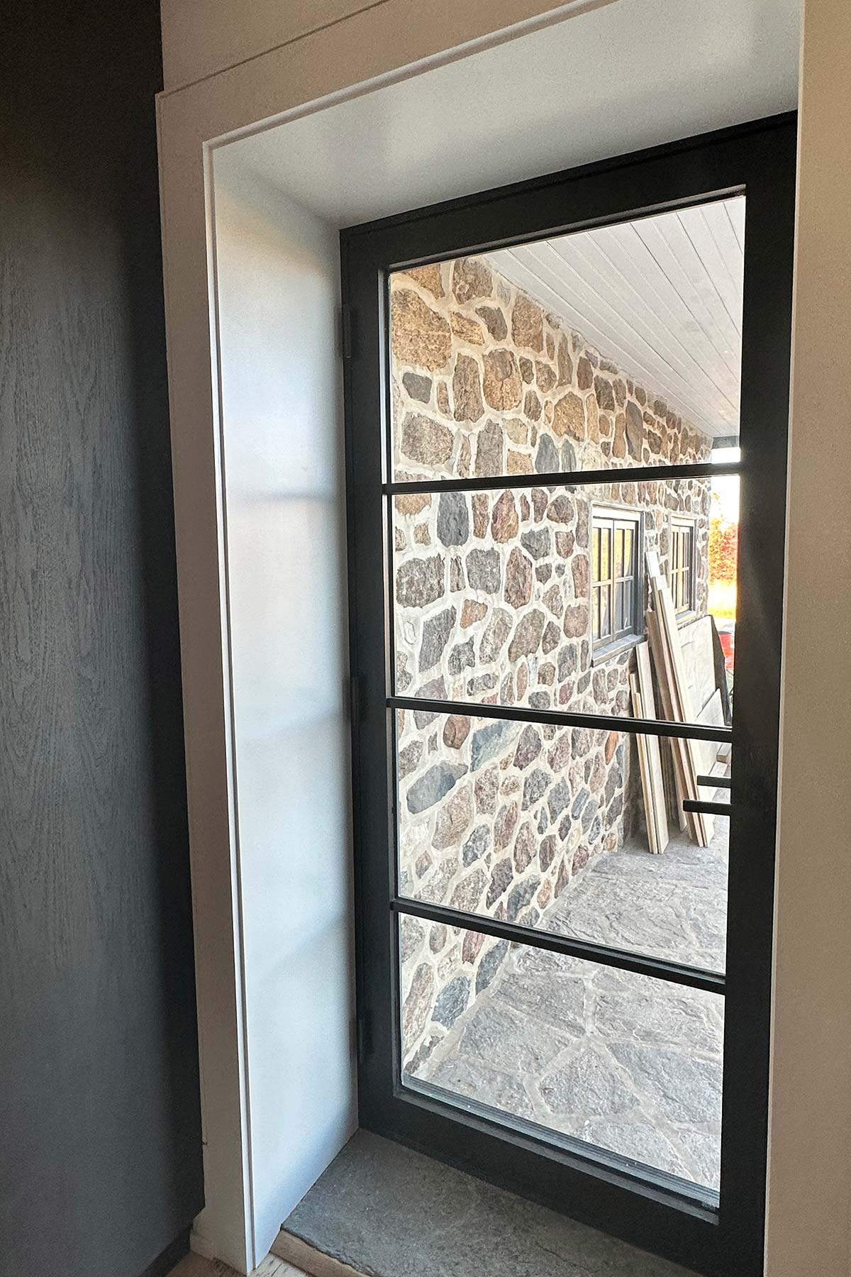 Black-framed glass door opens to stone-tiled patio with stone wall, two windows, and stacked wooden planks under a white-paneled ceiling.