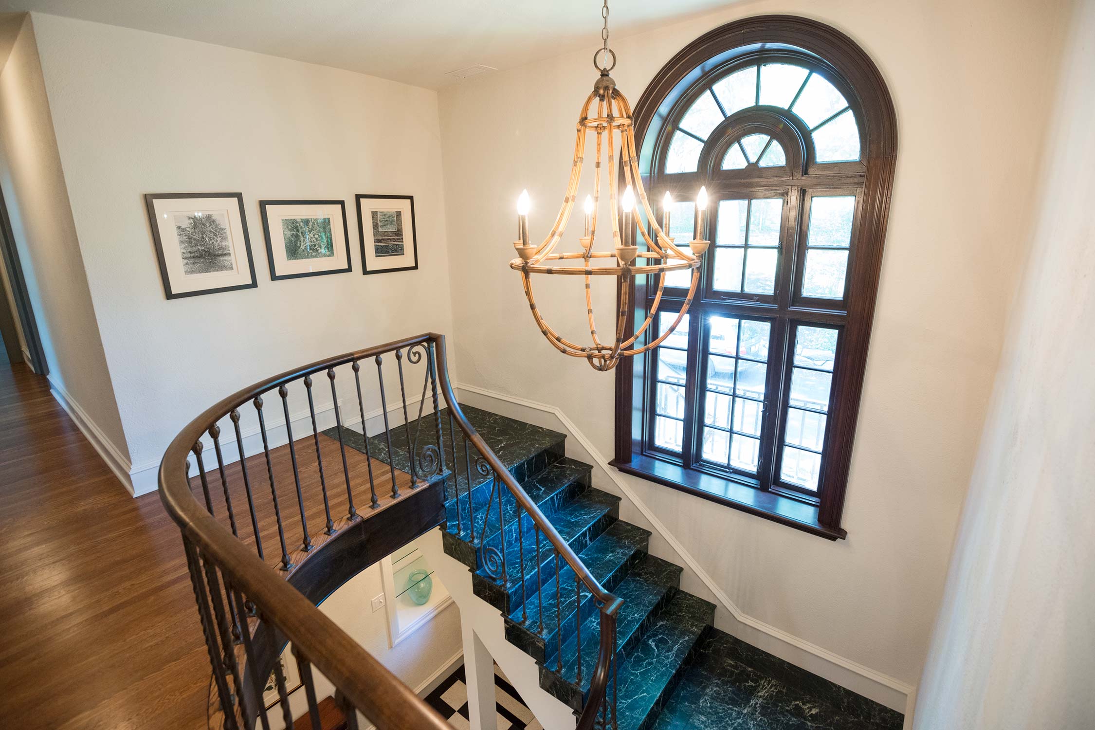 Elegant curved staircase featuring green marble steps, wooden handrail, bamboo chandelier, arched window, and framed wall art.