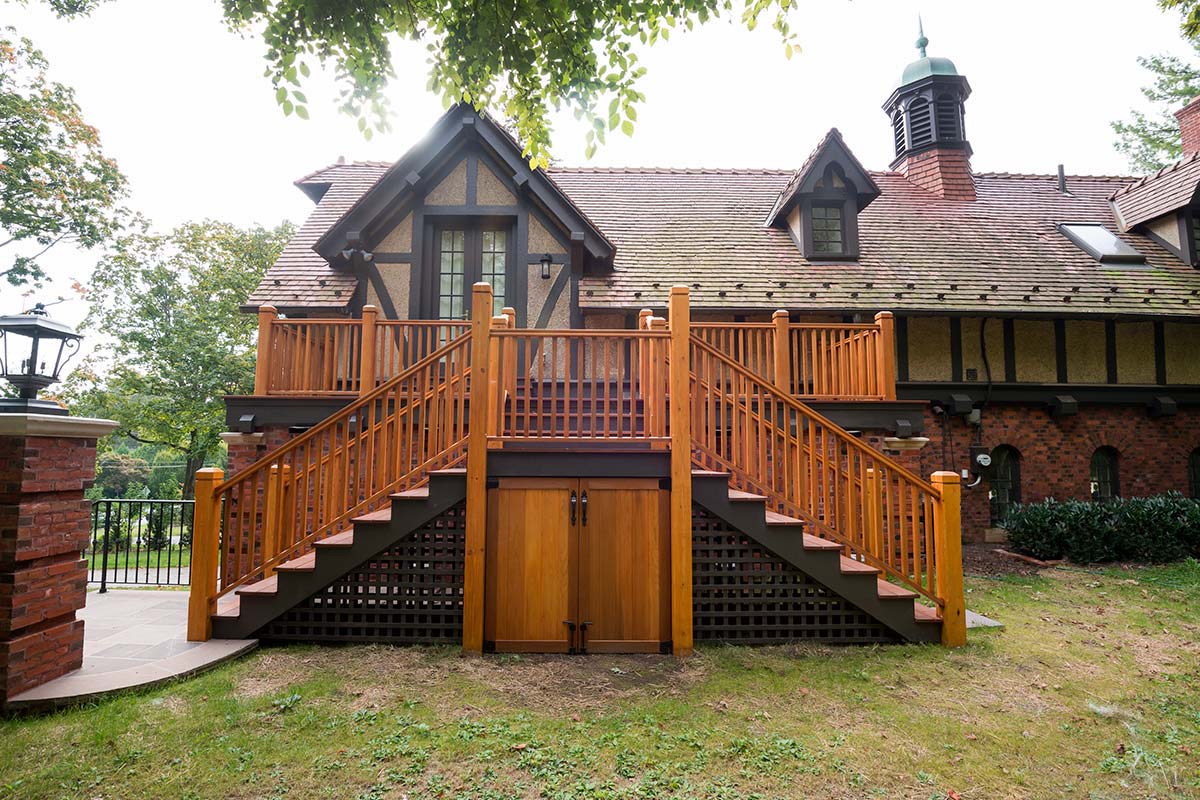 A brick and timber house with a wooden staircase, balcony, and double doors, set in a lawn with trees.