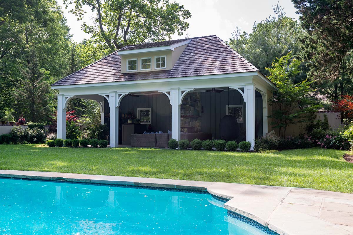 Charming poolside cabana with sloped roof and dormer windows. Features white columns, green lawn, surrounding trees, and a clear blue pool.