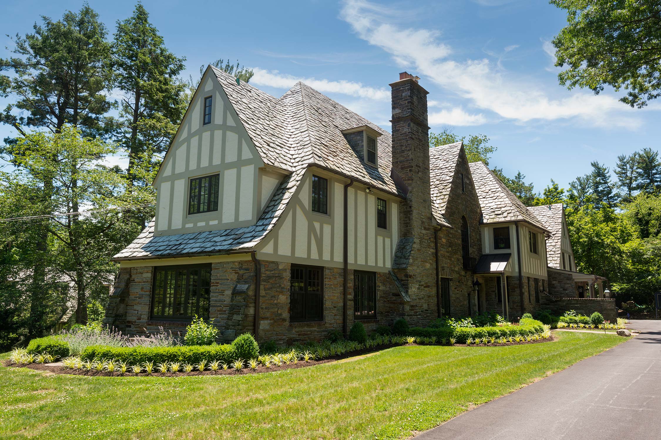Tudor-style stone house with steep shingle roof, chimney, and white trim, surrounded by green lawn and trees.