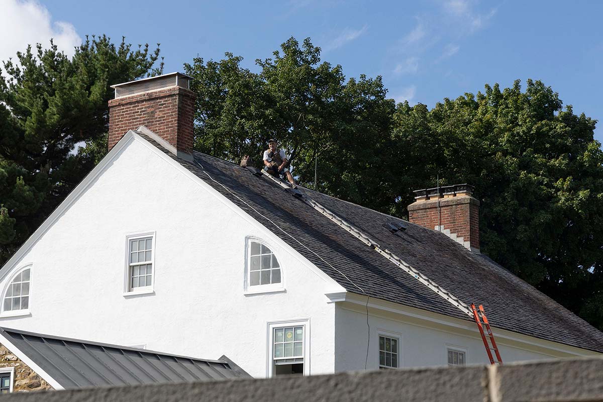 Person on ladder fixing roof of white house with black shingles, brick chimneys, and trees.