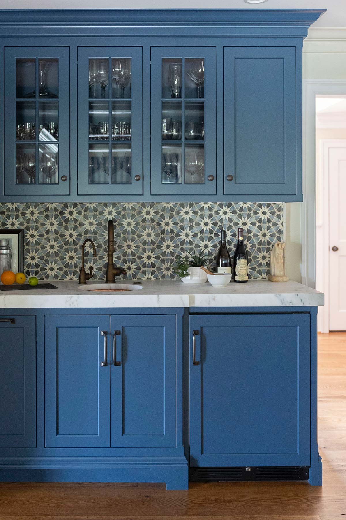 Blue kitchen cabinets with glass doors, star-patterned tile backsplash, and marble countertop. Includes sink, wine bottles, and fruit.