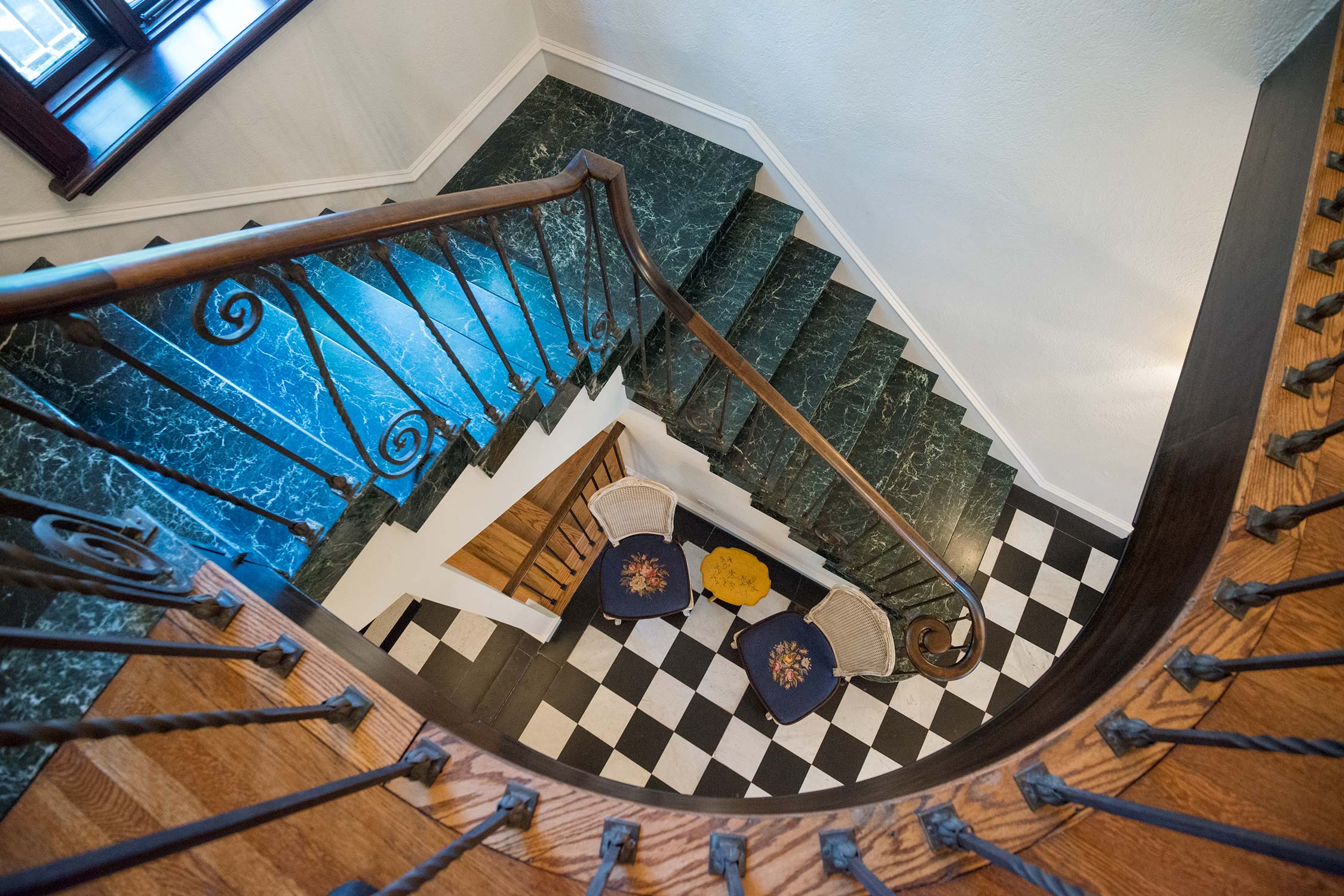 Elegant spiral staircase with green marble steps, wooden handrail, and wrought iron balusters, leading to a black-and-white checkered floor.