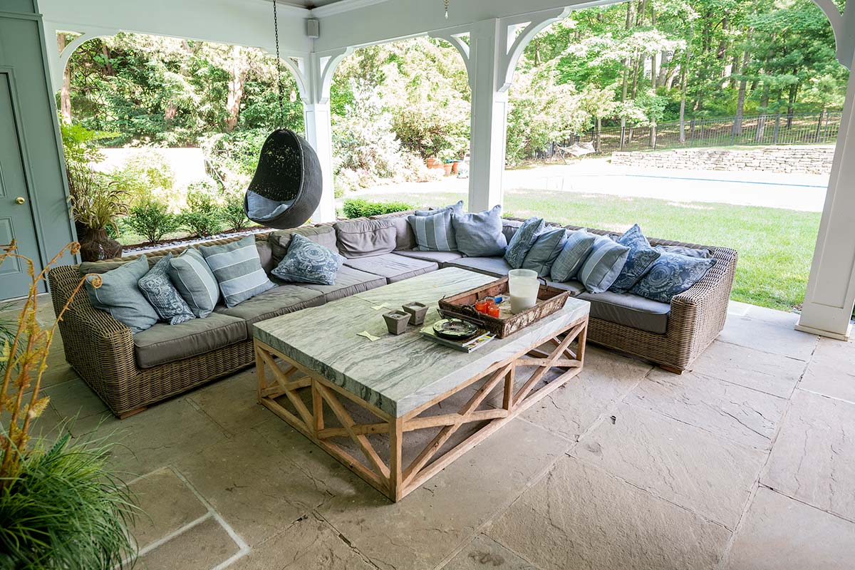 Cozy outdoor patio with wicker sectional sofa, marble coffee table, and hanging chair. Features stone flooring, lush greenery, and arched white columns.