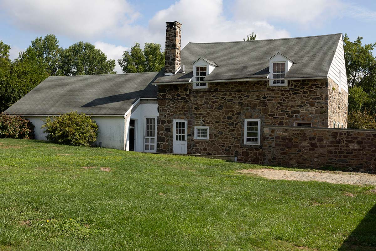 Stone house with white addition, black roof, dormer windows, and chimney, surrounded by green lawn and trees.