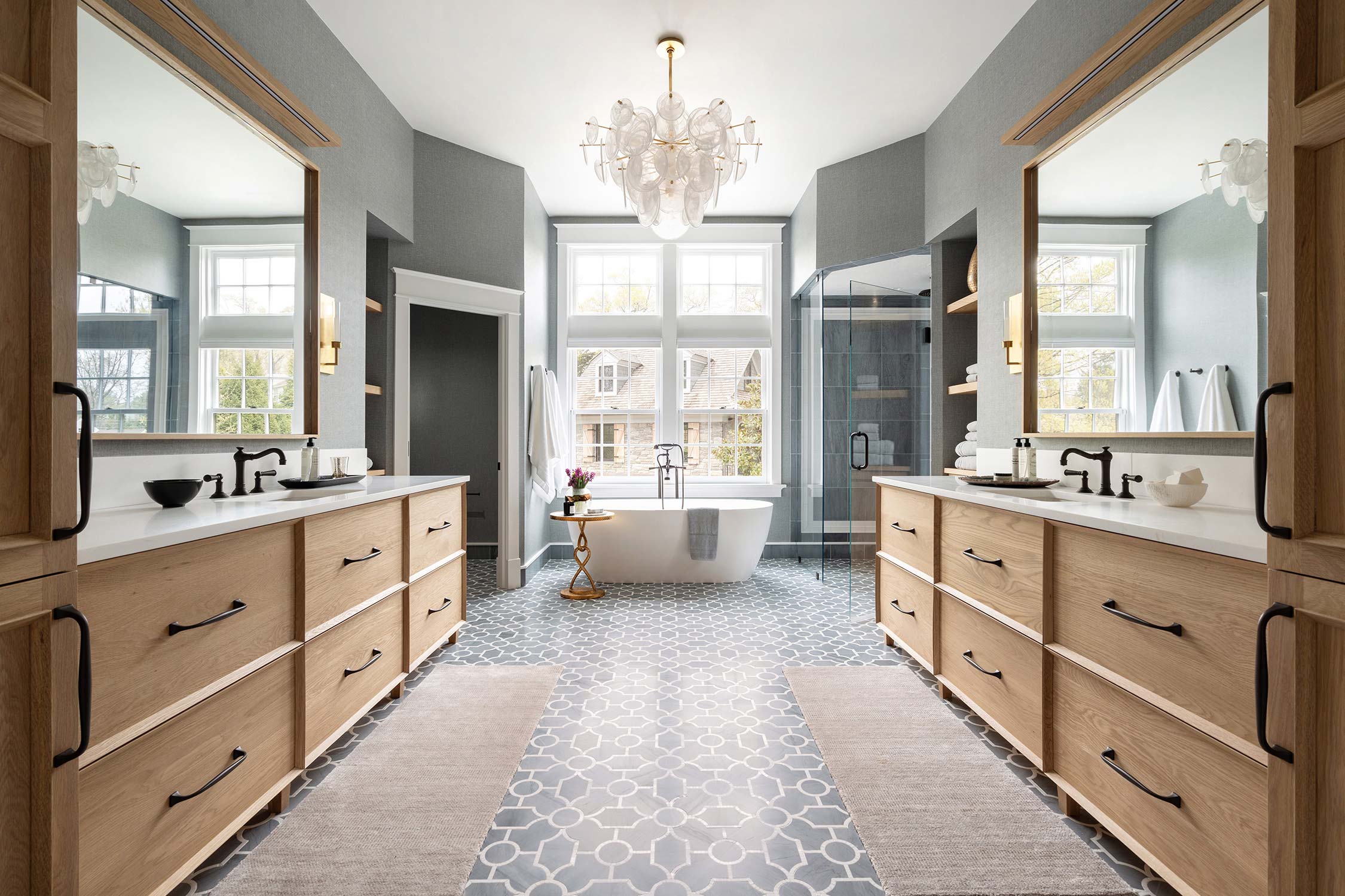 Luxurious bathroom with dual wooden vanities, a freestanding tub, large windows, and a chandelier, featuring patterned tile floors.