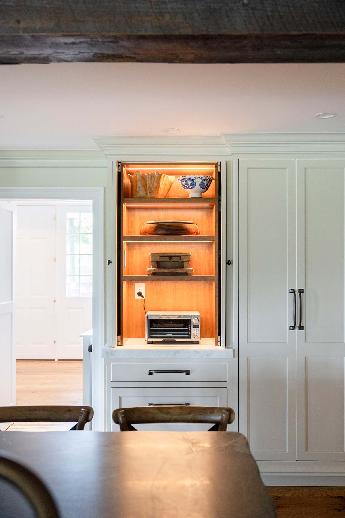 Open kitchen cabinet with lit shelves holding a toaster oven, bowls, and bread. White cabinets and wooden chairs surround a dark table on wood floor.