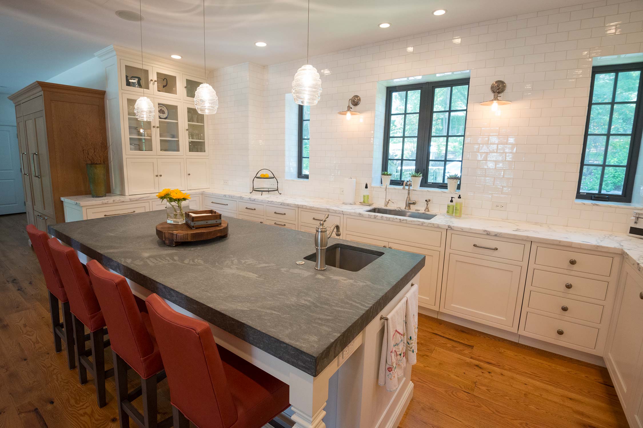 Bright kitchen with white cabinets, subway tile backsplash, island, red bar stools, and large windows.