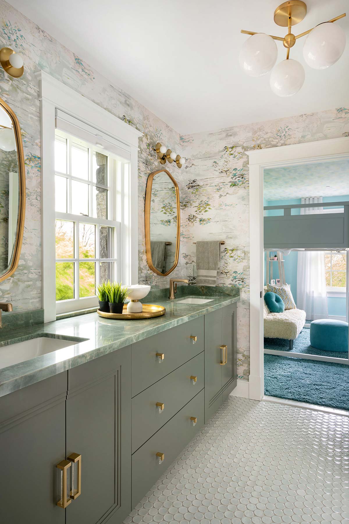 Modern bathroom with dual gray vanities, gold accents, large mirrors, and a window, featuring white penny tile floor and floral wallpaper.