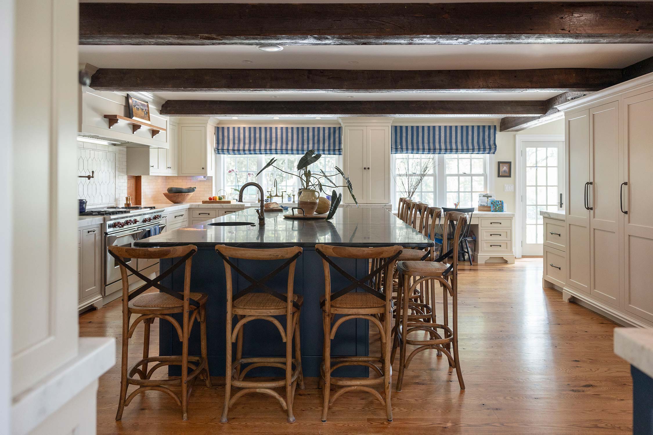 Spacious kitchen with blue island, wooden chairs, and white cabinets. Features striped curtains, wood beams, and large windows over wood floor.