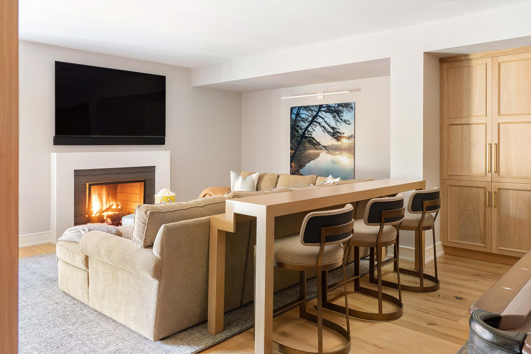 Cozy living room with a beige sofa, fireplace, flat-screen TV, and a wooden counter with bar stools, featuring a nature painting.