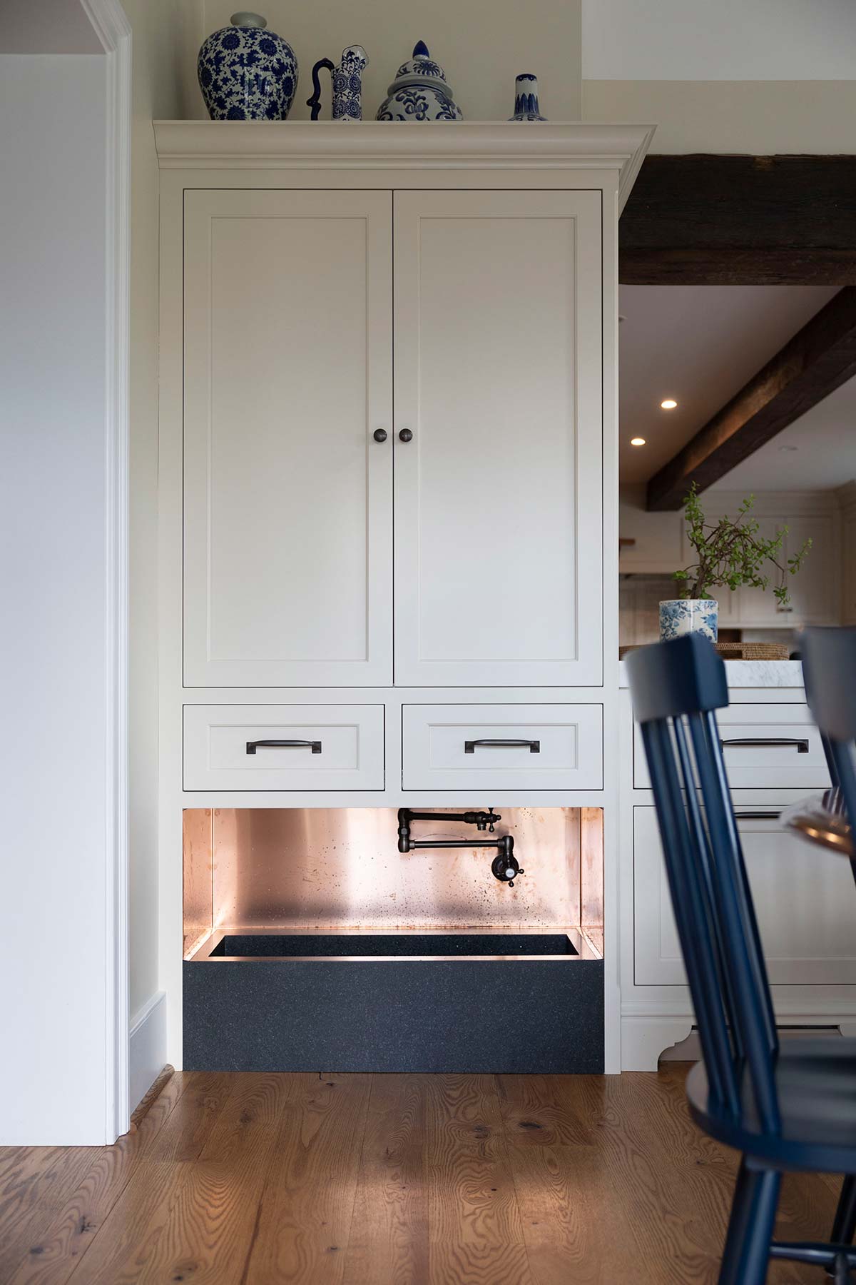 White cabinet with blue decor items on top, featuring a copper sink and faucet. Built-in dog bed under cabinet, with wooden chair and floor.