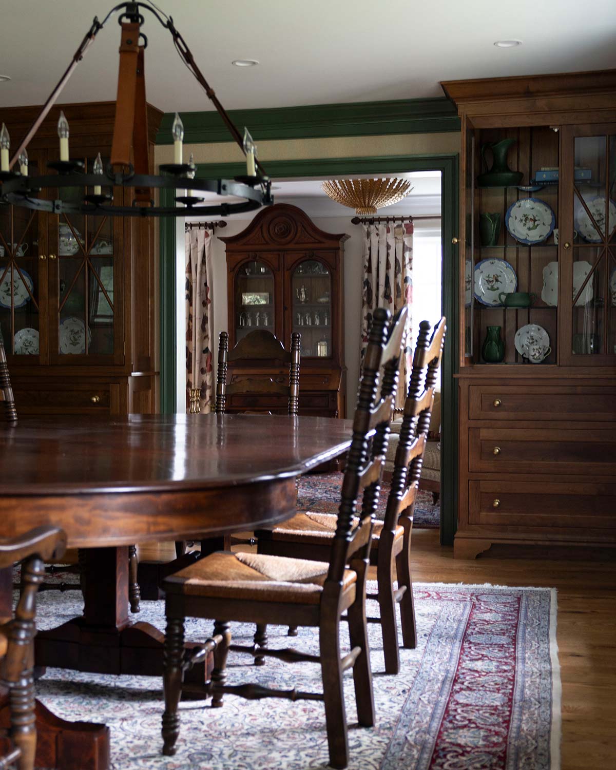 Elegant dining room with a polished wooden table, rustic chairs, a chandelier, and cabinets displaying decorative plates, accented by green wainscoting.
