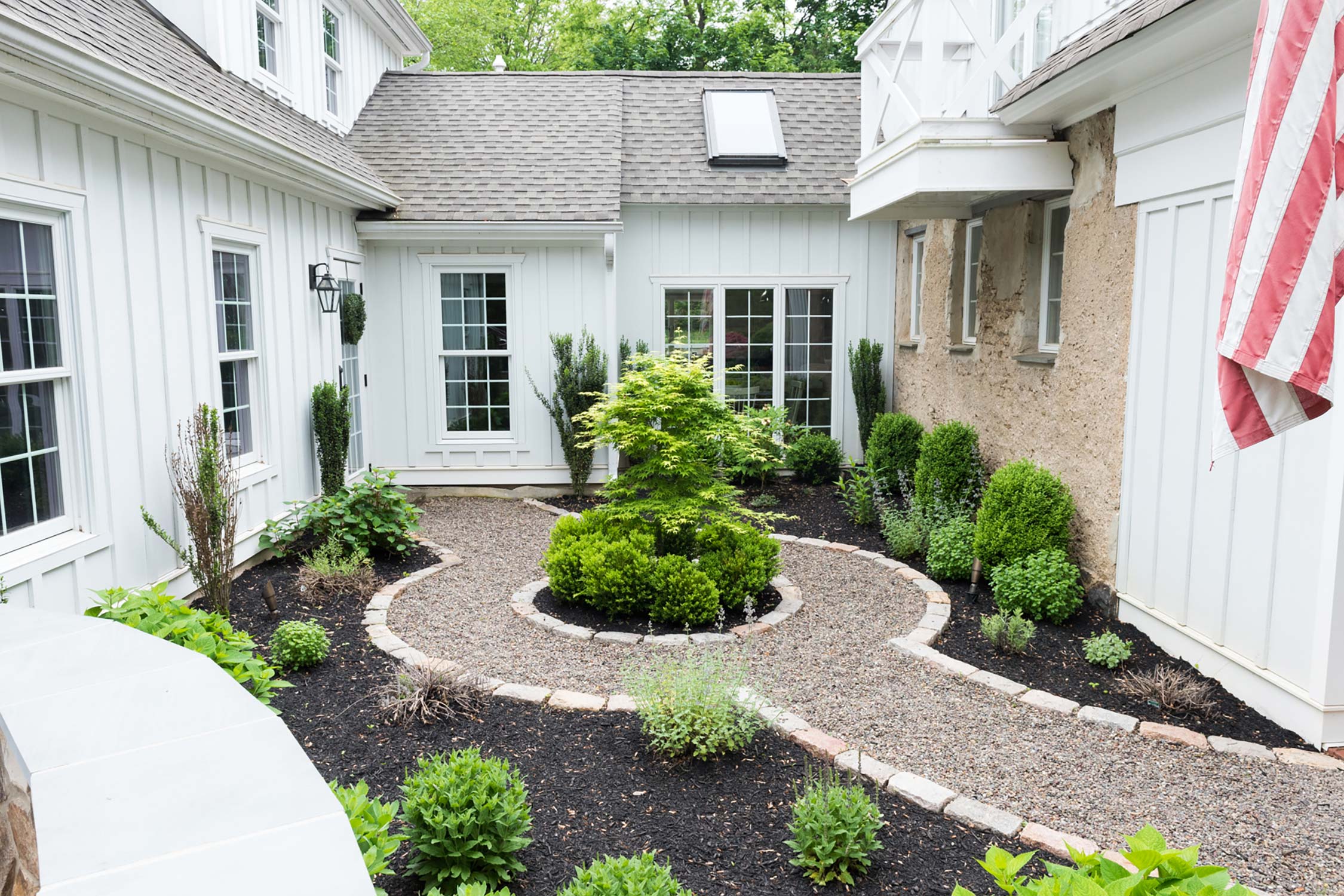 A courtyard with a gravel path, circular garden bed, shrubs, and a flag, surrounded by white and stone buildings.