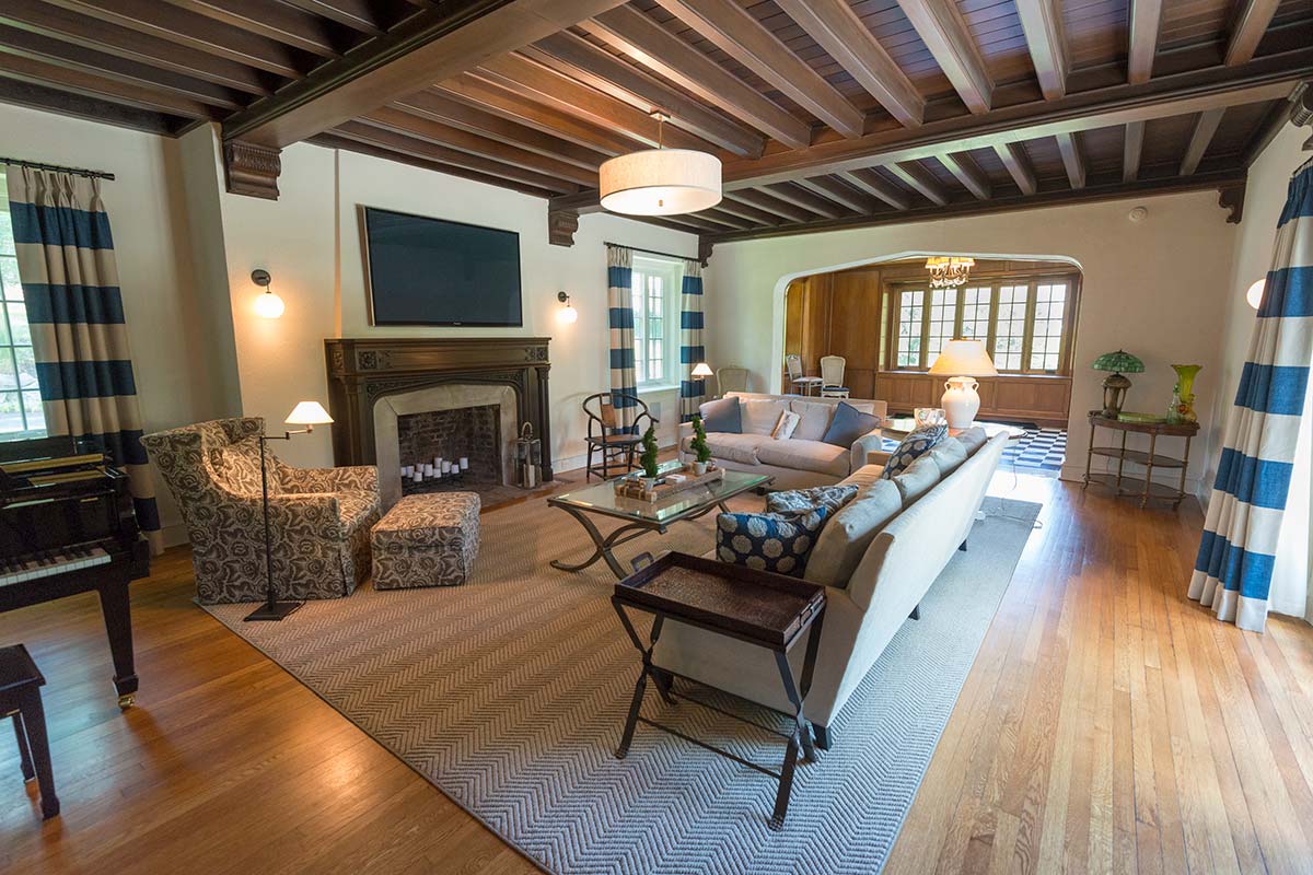 Elegant living room with wooden beams, fireplace, TV, light sofa, patterned chair, and piano, with blue-striped curtains.