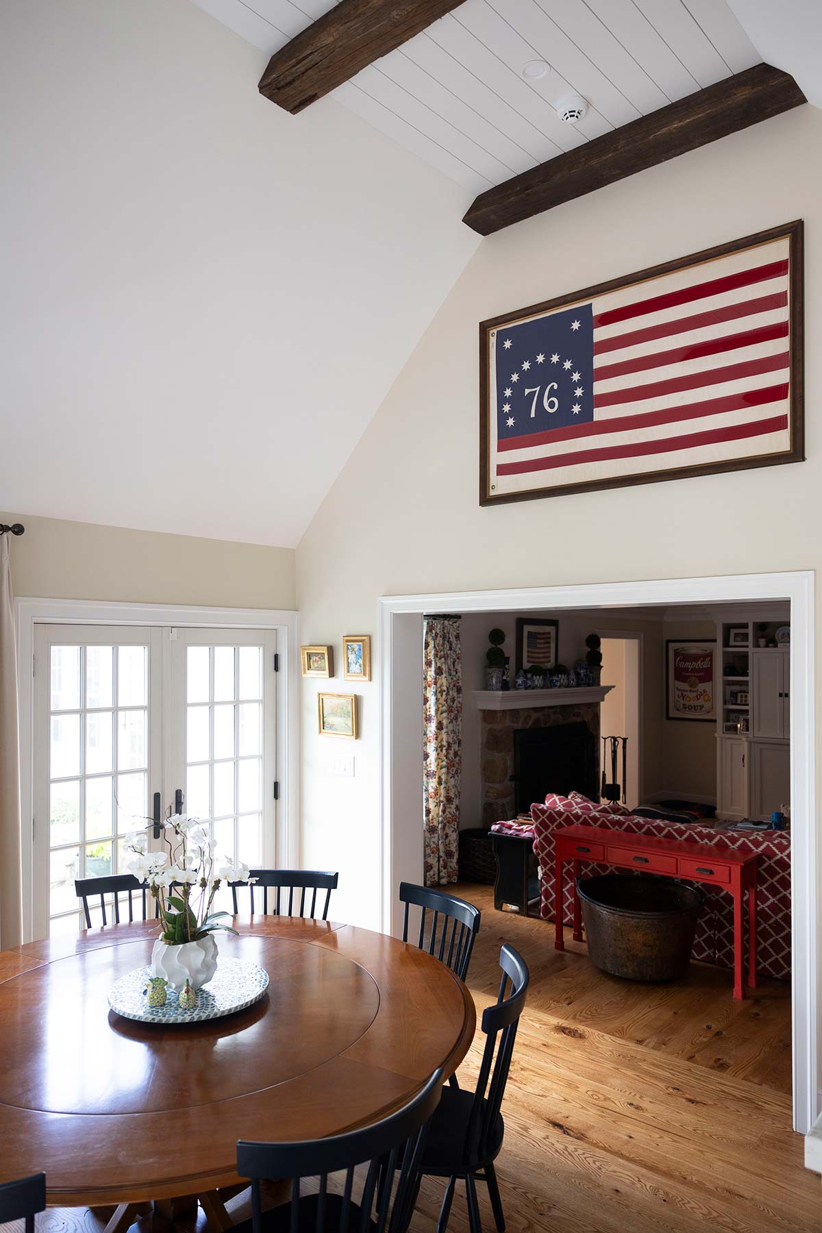 Dining area with round wooden table, black chairs, and white flowers. Vintage 76 flag on wall, wood beams, and red table visible through doorway.