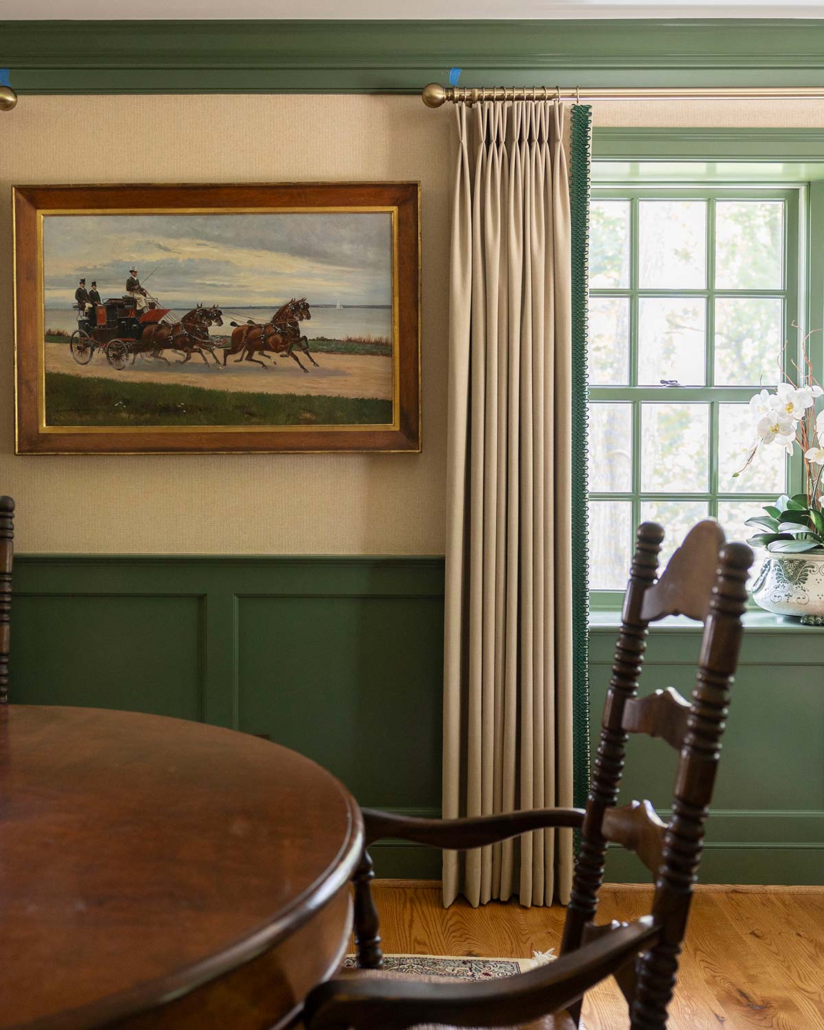 Charming dining area with a wooden table, rustic chair, and a painting of a horse-drawn carriage, accented by green wainscoting and a window with curtains.