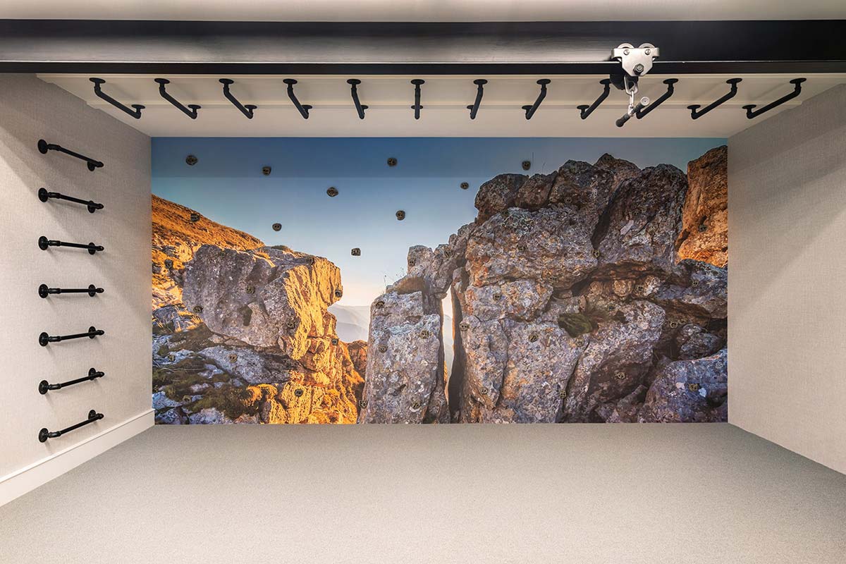 Indoor climbing wall with a scenic rocky canyon mural, equipped with black metal handholds and a pulley system on the ceiling.