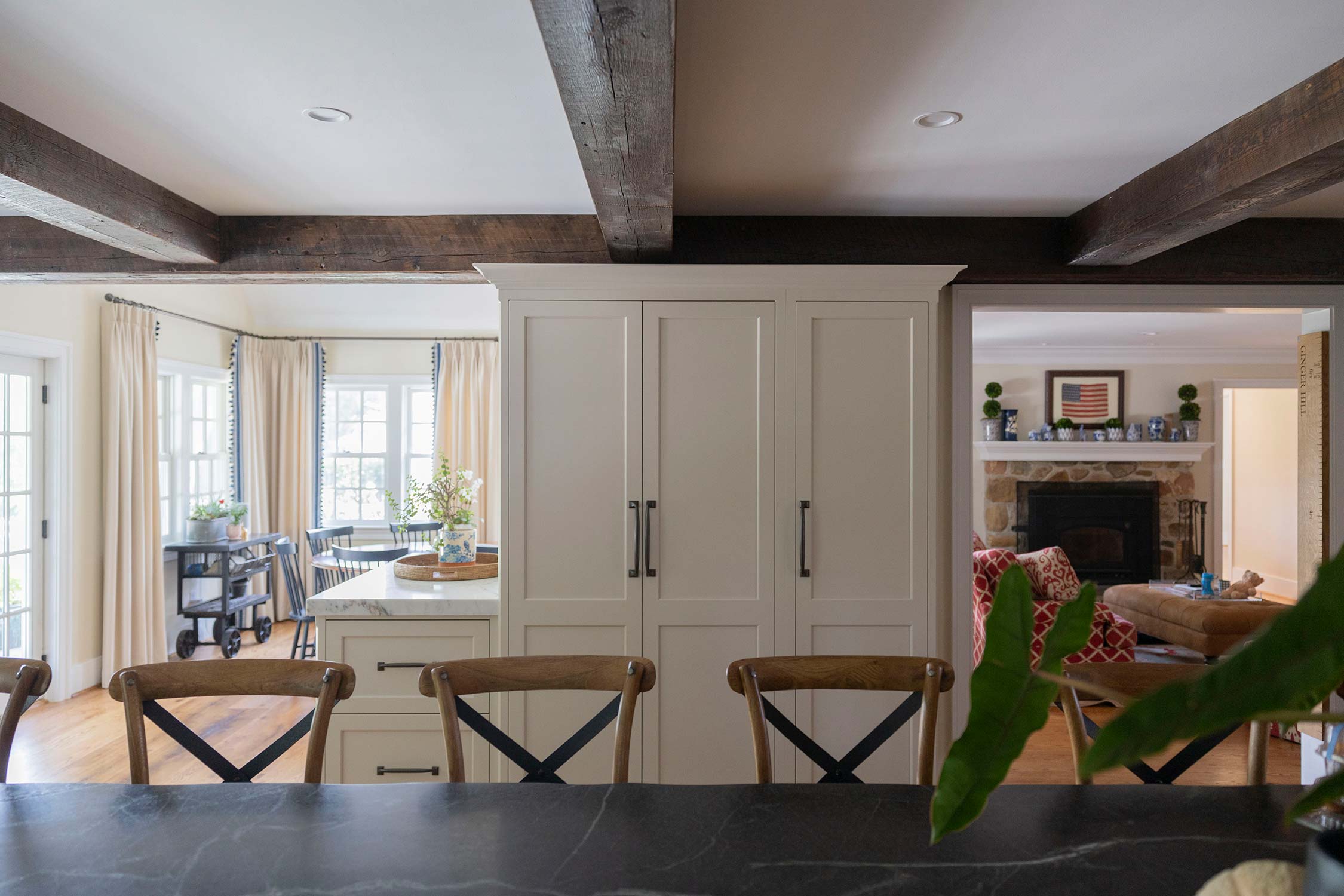 Modern kitchen with white cabinets, wooden chairs, and dark countertop. Features wood beams, large windows, and a fireplace visible in the background.