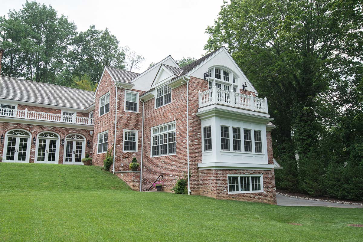 A brick house with large windows, a balcony, and a well-maintained lawn, surrounded by dense trees.