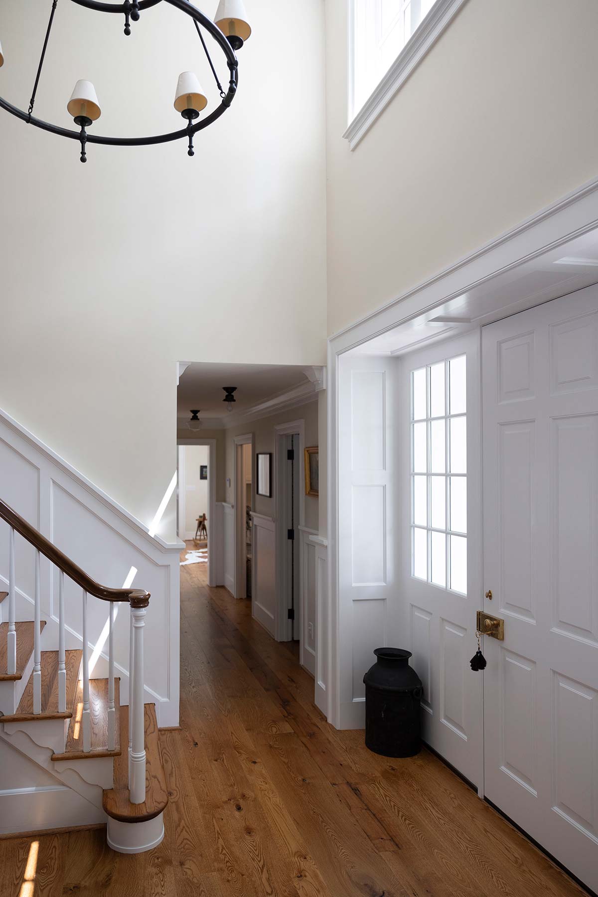 Bright foyer with a white staircase, wooden floors, a chandelier, and a white door with a black bucket, leading to a hallway.