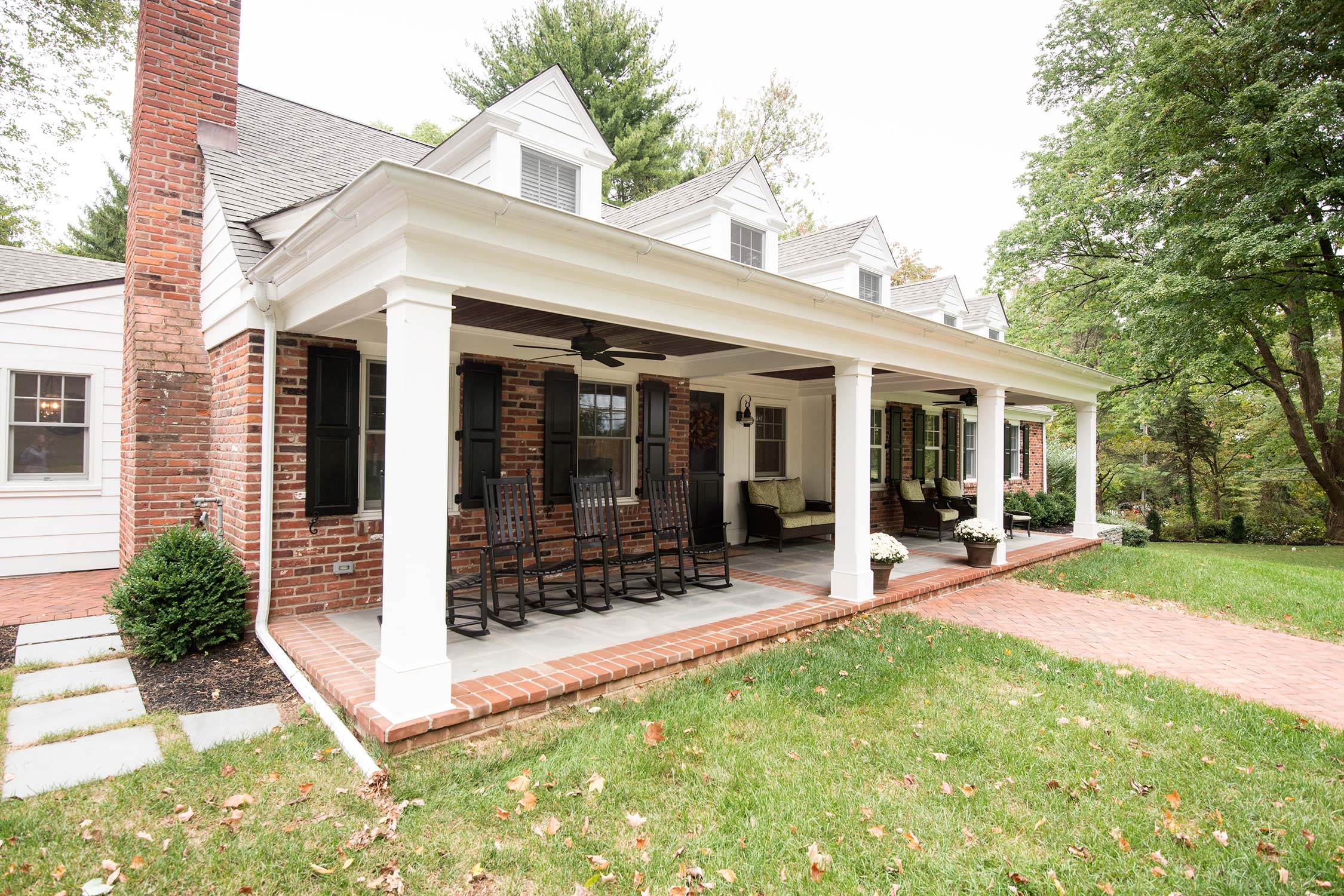 A brick and white house with a covered porch, rocking chairs, and a brick pathway, surrounded by a lawn and trees.