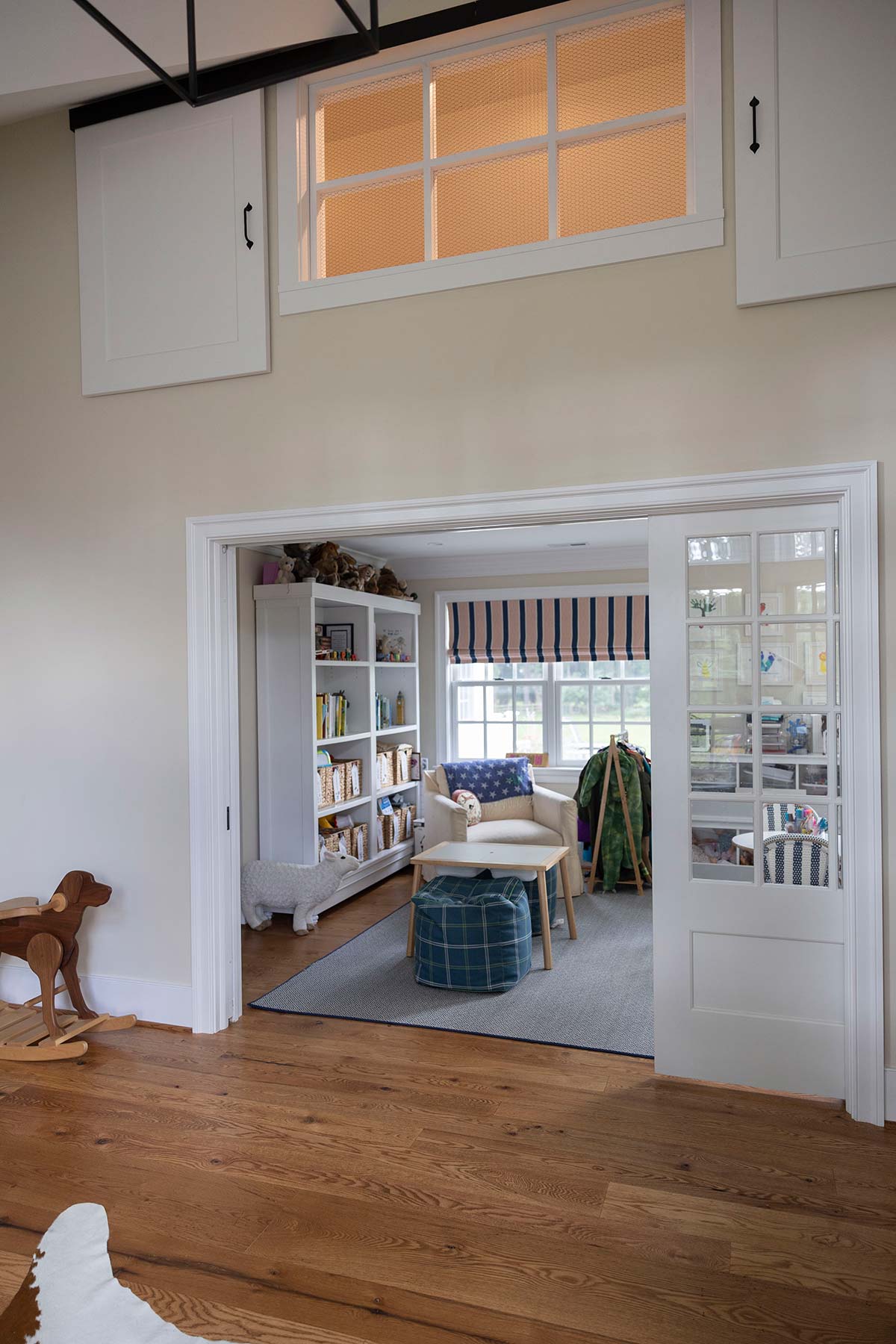 A cozy children's room with wooden floors, a rocking horse, bookshelves, and a play area, featuring natural light through large windows and sliding doors.
