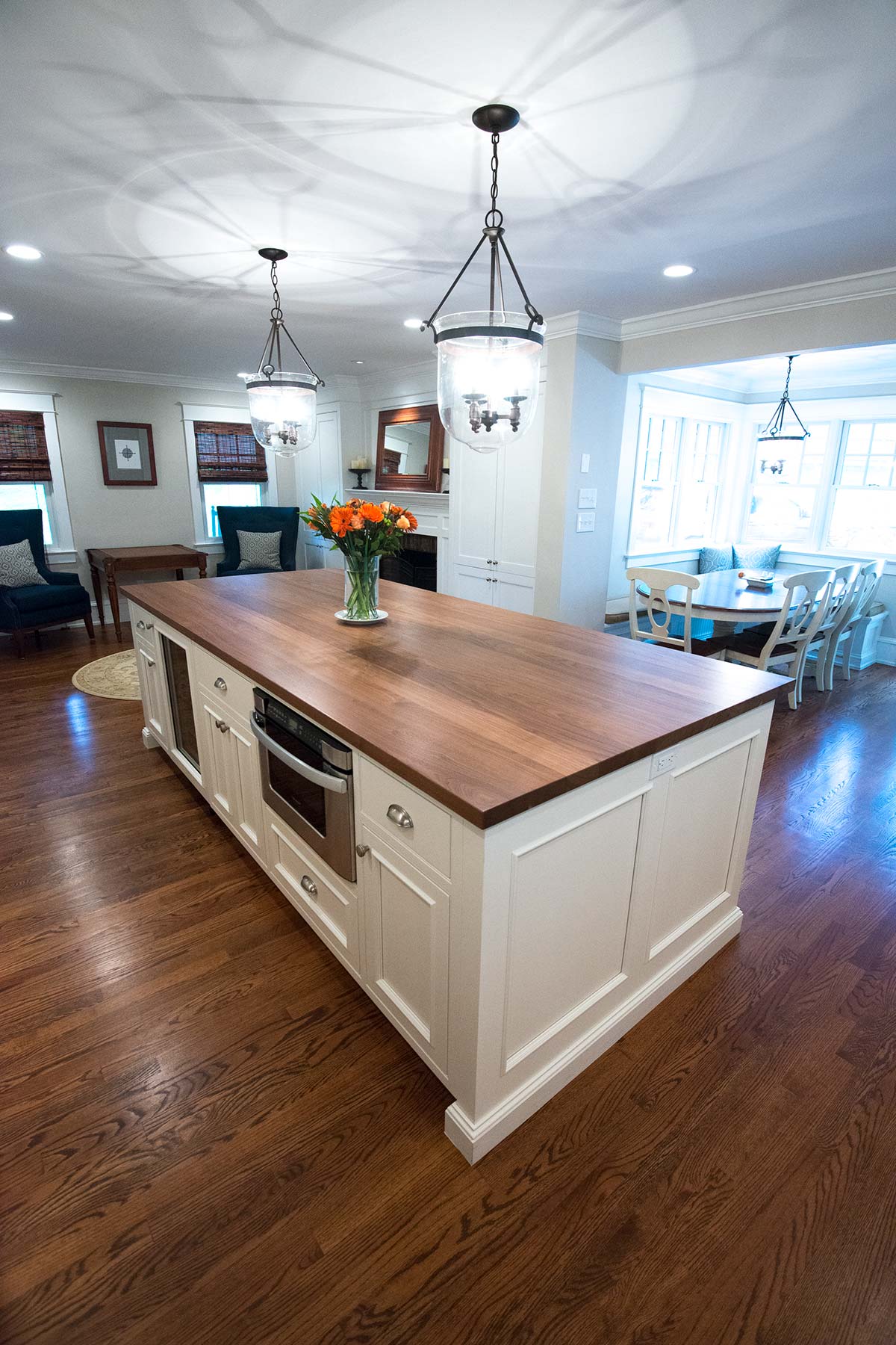 A kitchen with a large island, wooden countertop, vase of flowers, and pendant lights, opening to a dining area.