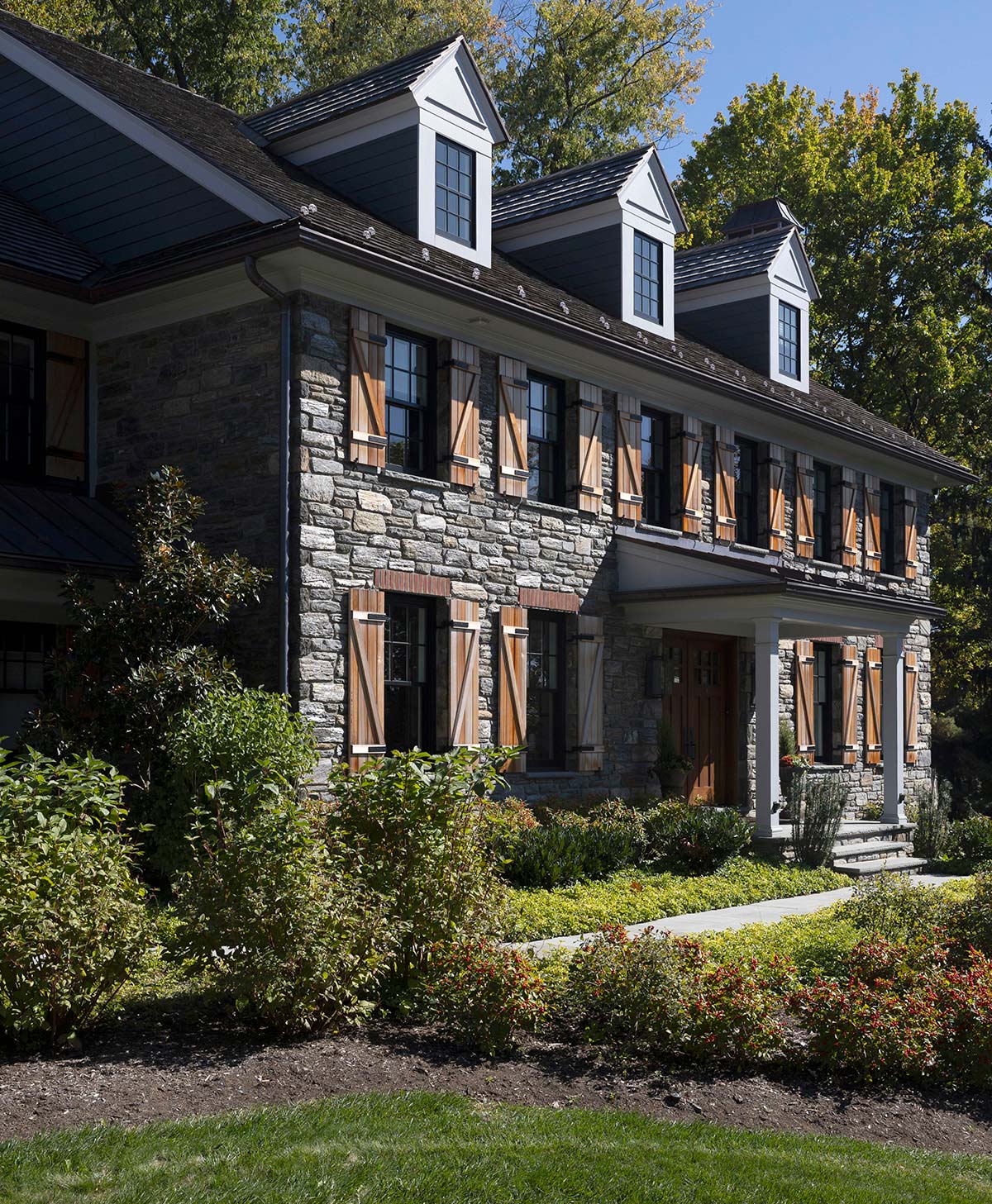 Stone house with dark roof, dormer windows, and wooden shutters, surrounded by lush greenery and a pathway leading to the entrance.