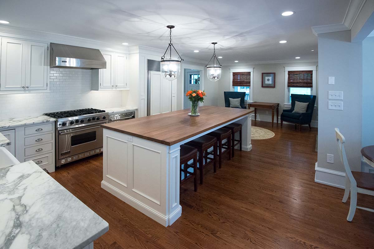A kitchen with a large island, wooden countertop, stools, vase of flowers, and pendant lights, opening to a sitting area.