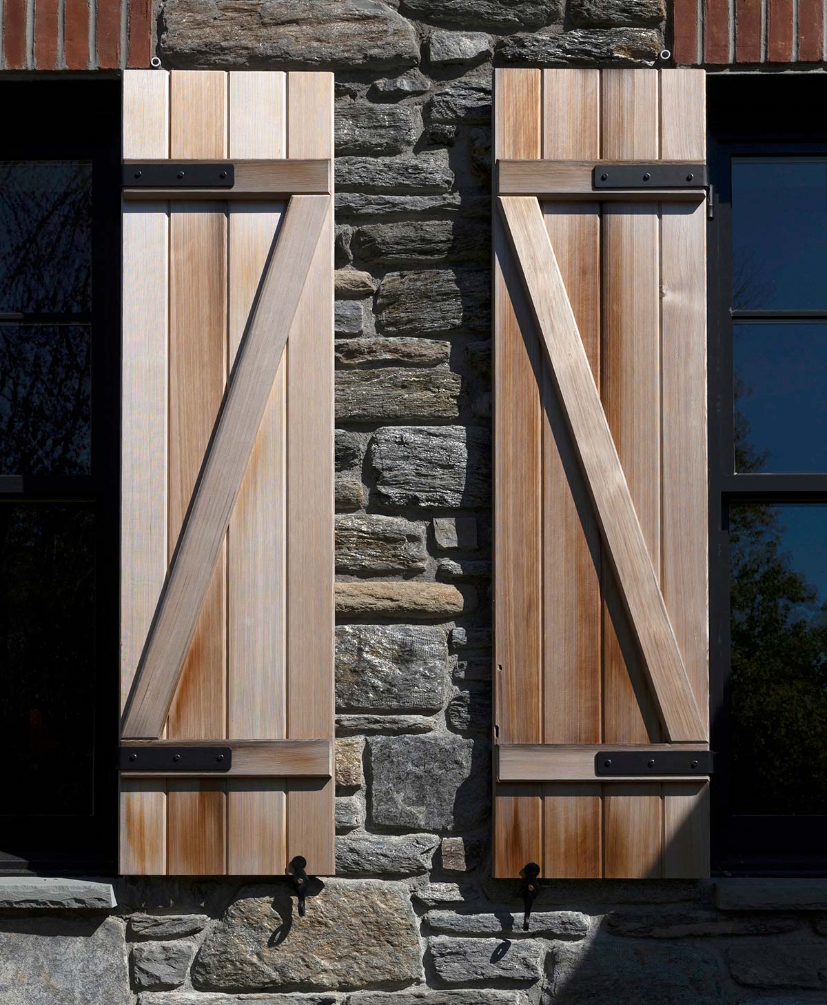 Wooden shutters with diagonal braces on a stone wall, next to a dark-framed window, showcasing rustic architectural detail.