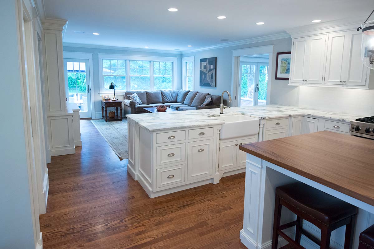 A modern kitchen with white cabinets, marble counters, and a wooden island, opening to a cozy living room with a gray sofa, rug, and large windows.
