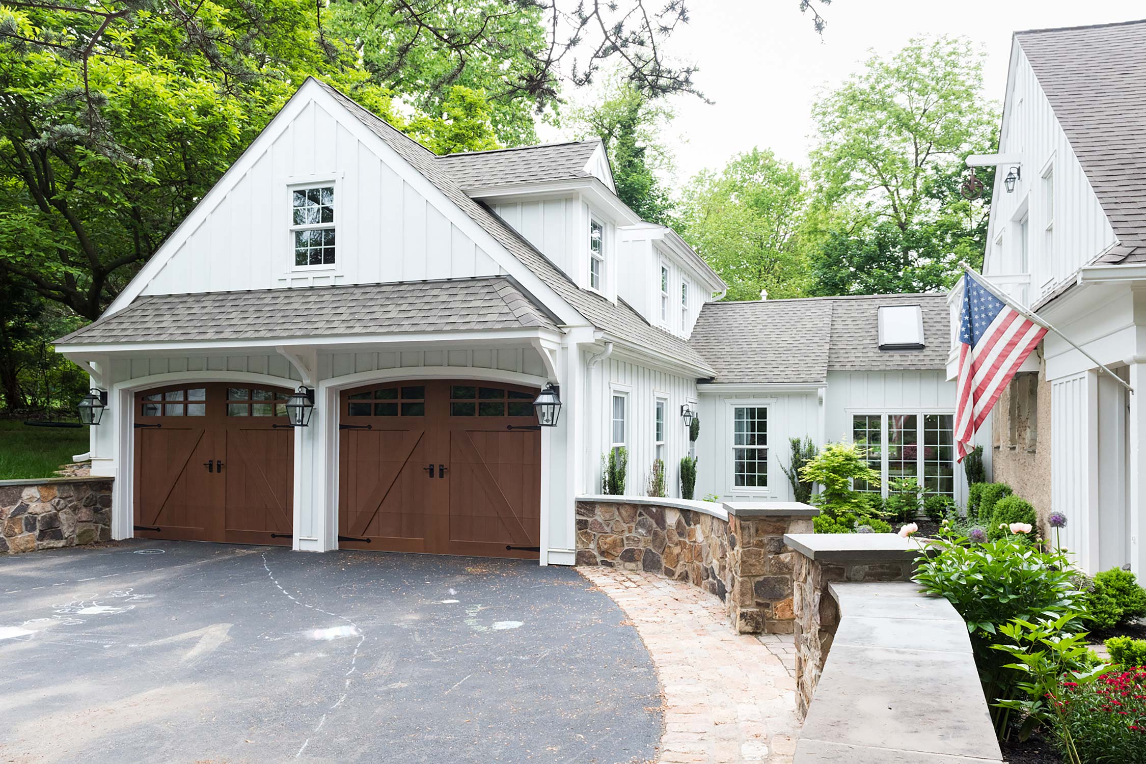 A white house with a gray roof, two wooden garage doors, stone wall, American flag, and lush greenery around.