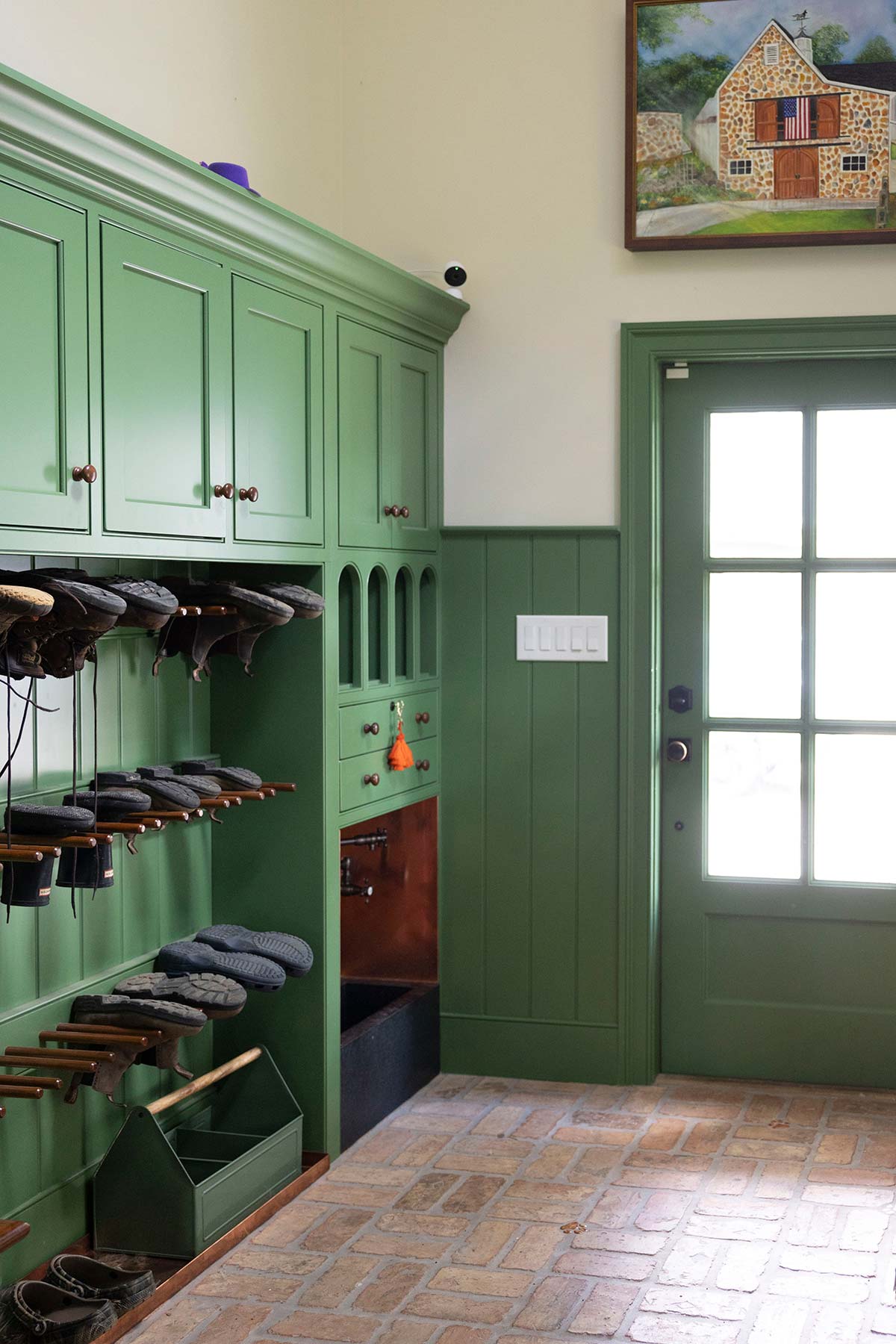 Green cabinetry with shoe racks and storage, featuring a painting of a house. Brick floor and a door with natural light enhance the entryway.