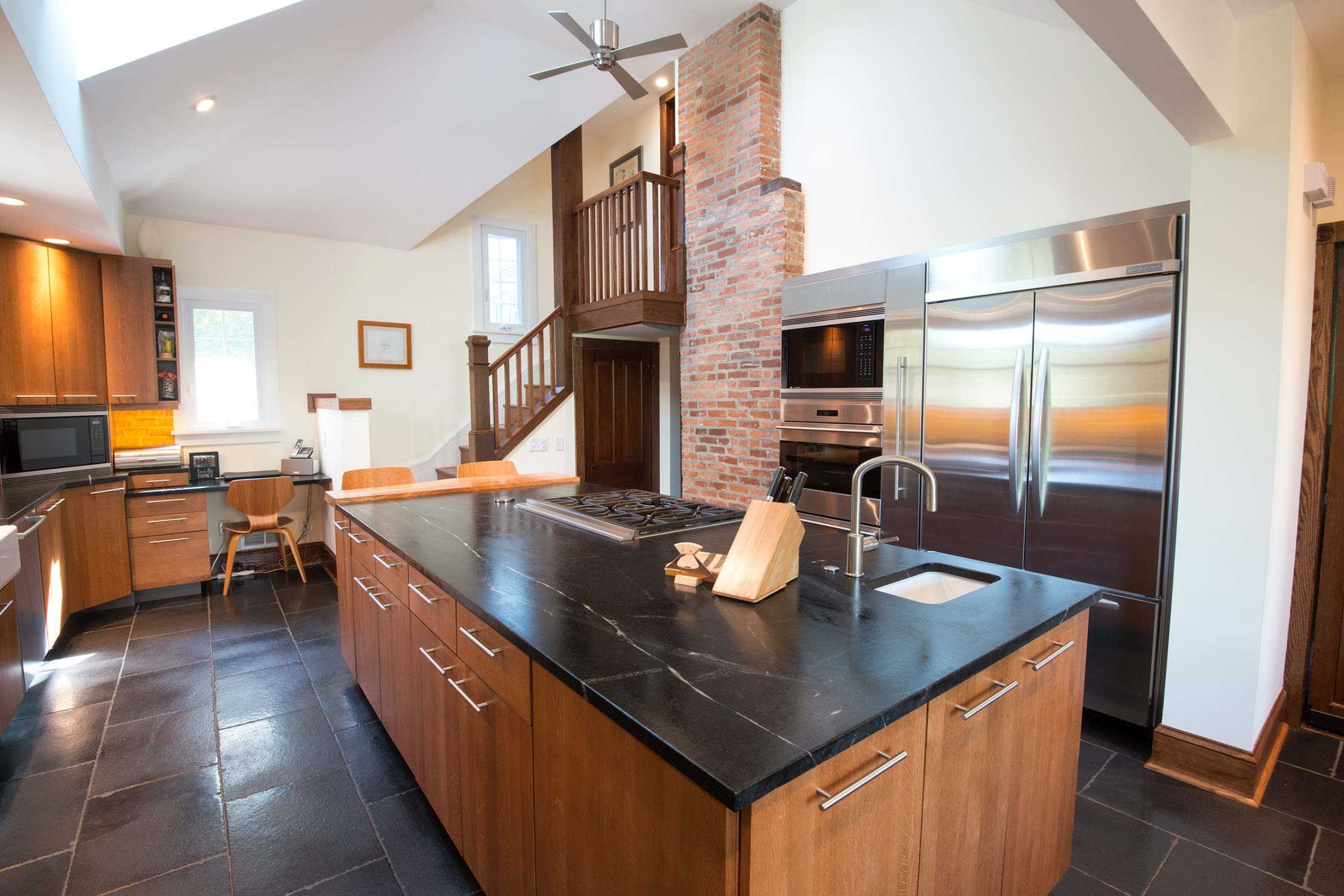 A spacious kitchen with wooden cabinets, black granite counters, stainless steel appliances, a brick accent wall, and a staircase leading to a loft.