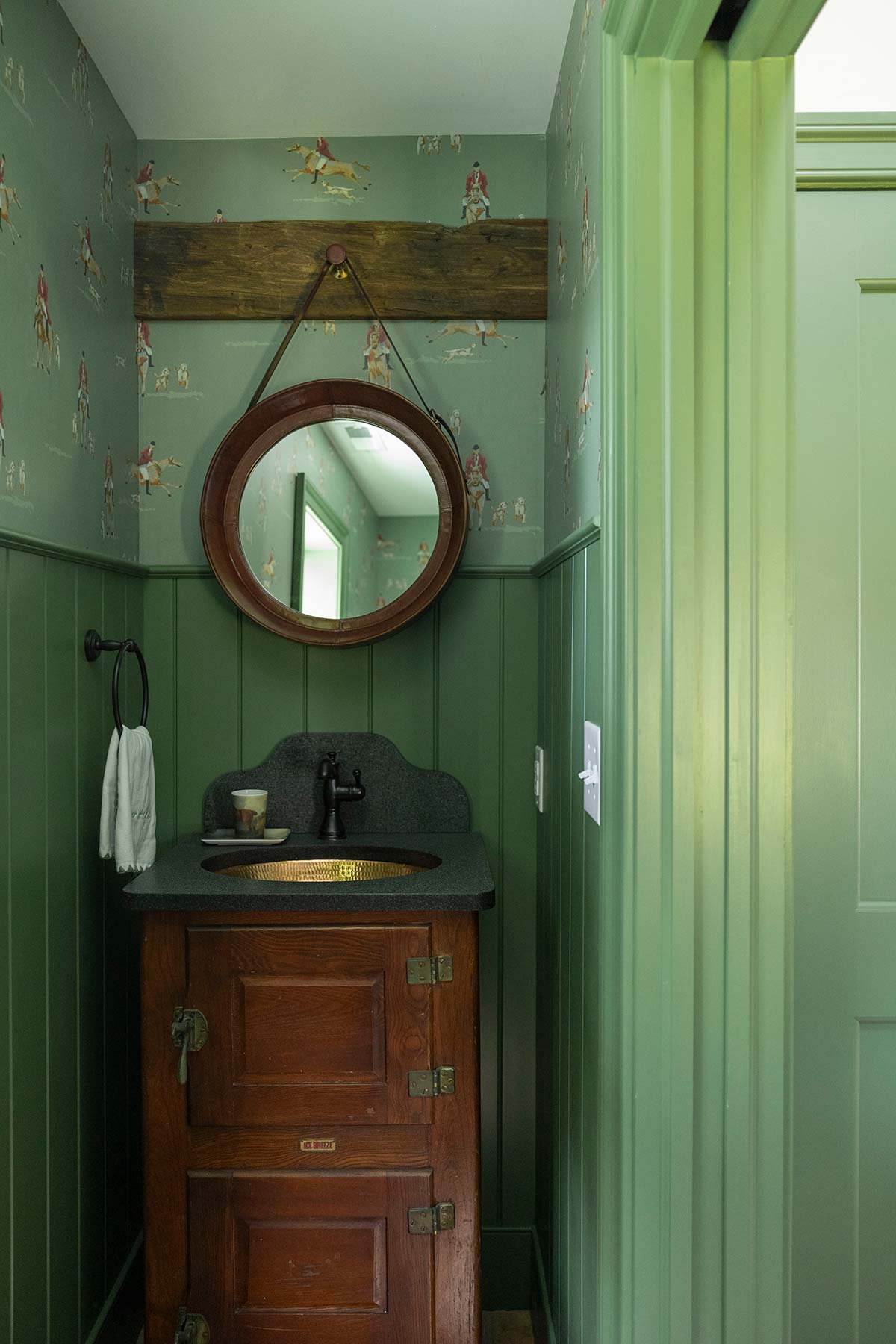 Small green bathroom with horse-themed wallpaper, round mirror, and wooden vanity with copper sink. Includes a towel and natural light.