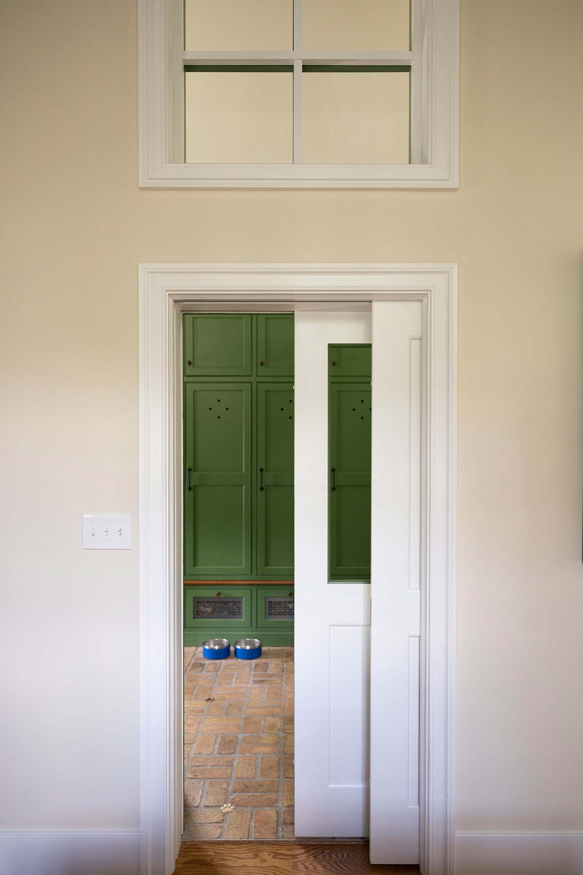 A cozy room with a pocket door revealing a green cabinet area, brick floor, and pet bowls, framed by a high window.