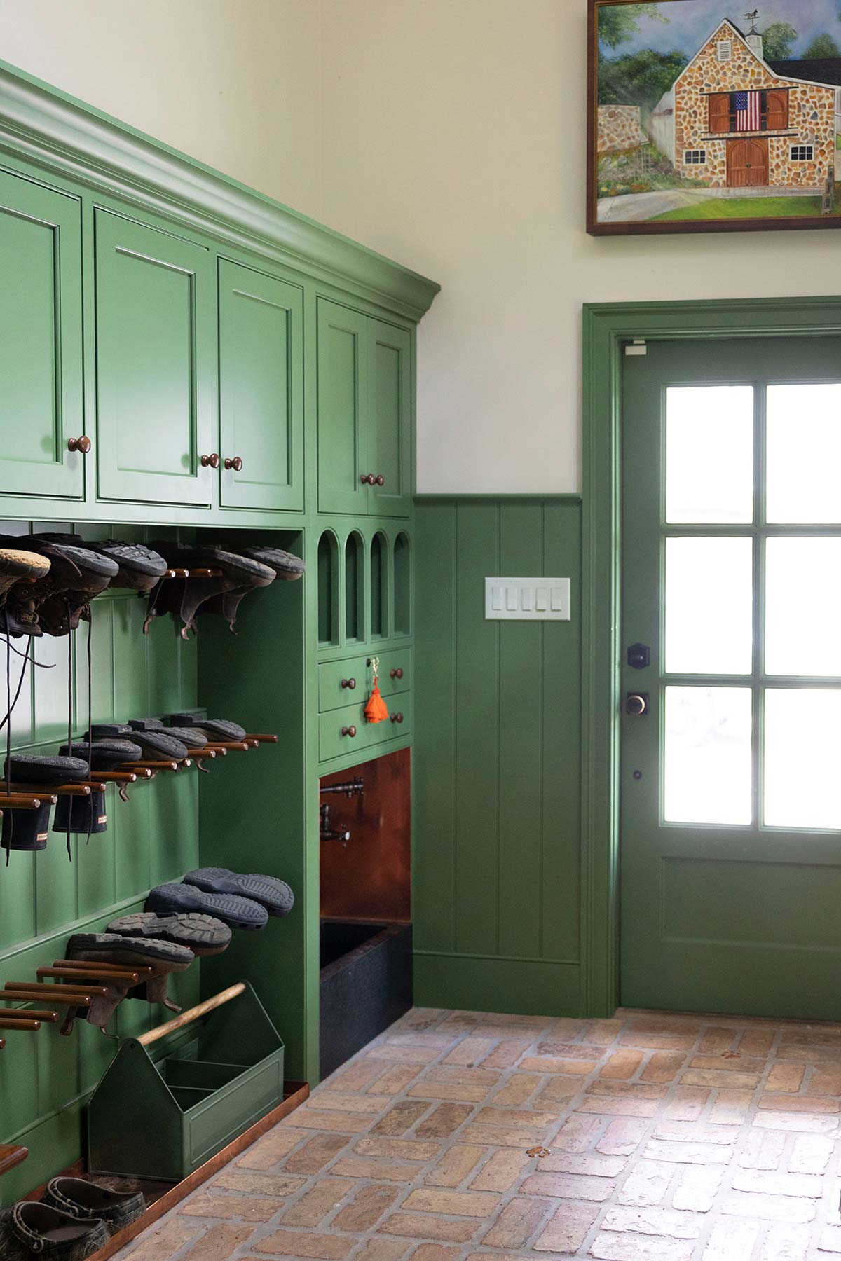 Green cabinetry with shoe racks and storage, featuring a painting of a house. Brick floor and a door with natural light enhance the entryway.