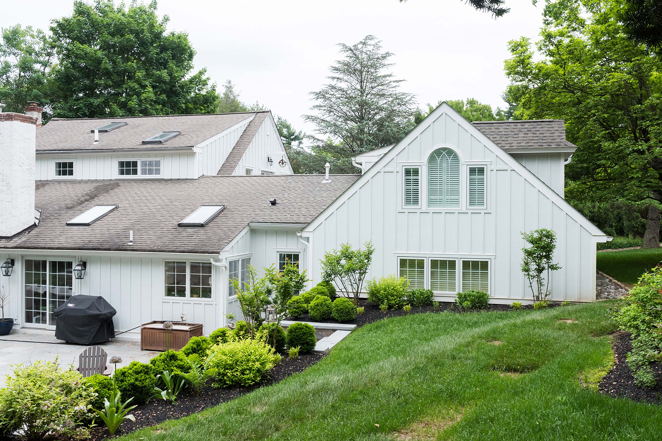 A white house with a gray roof, large windows, a covered patio with a grill, and a landscaped yard with shrubs and a lawn.