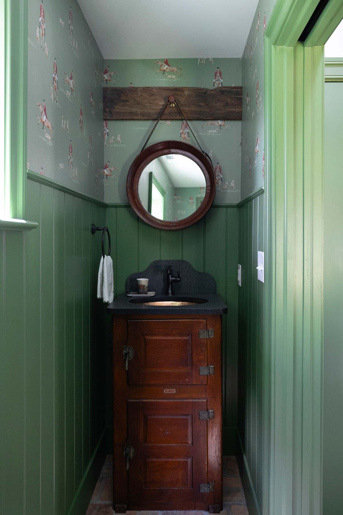 Small green bathroom with horse-themed wallpaper, round mirror, and wooden vanity with copper sink. Includes a towel and natural light.