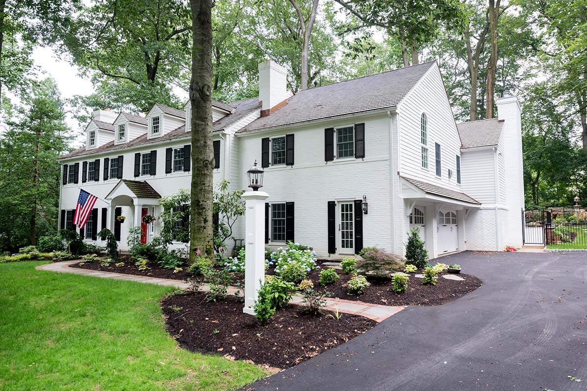 A large white house with black shutters, a red door, American flag, and well-maintained landscaping with a stone path and driveway.