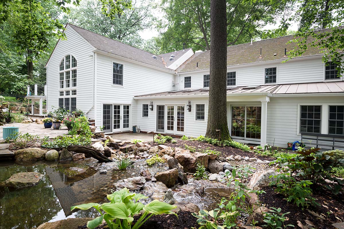 A white house with large windows, a patio, and a backyard featuring a small pond with rocks, plants, and a flowing stream.
