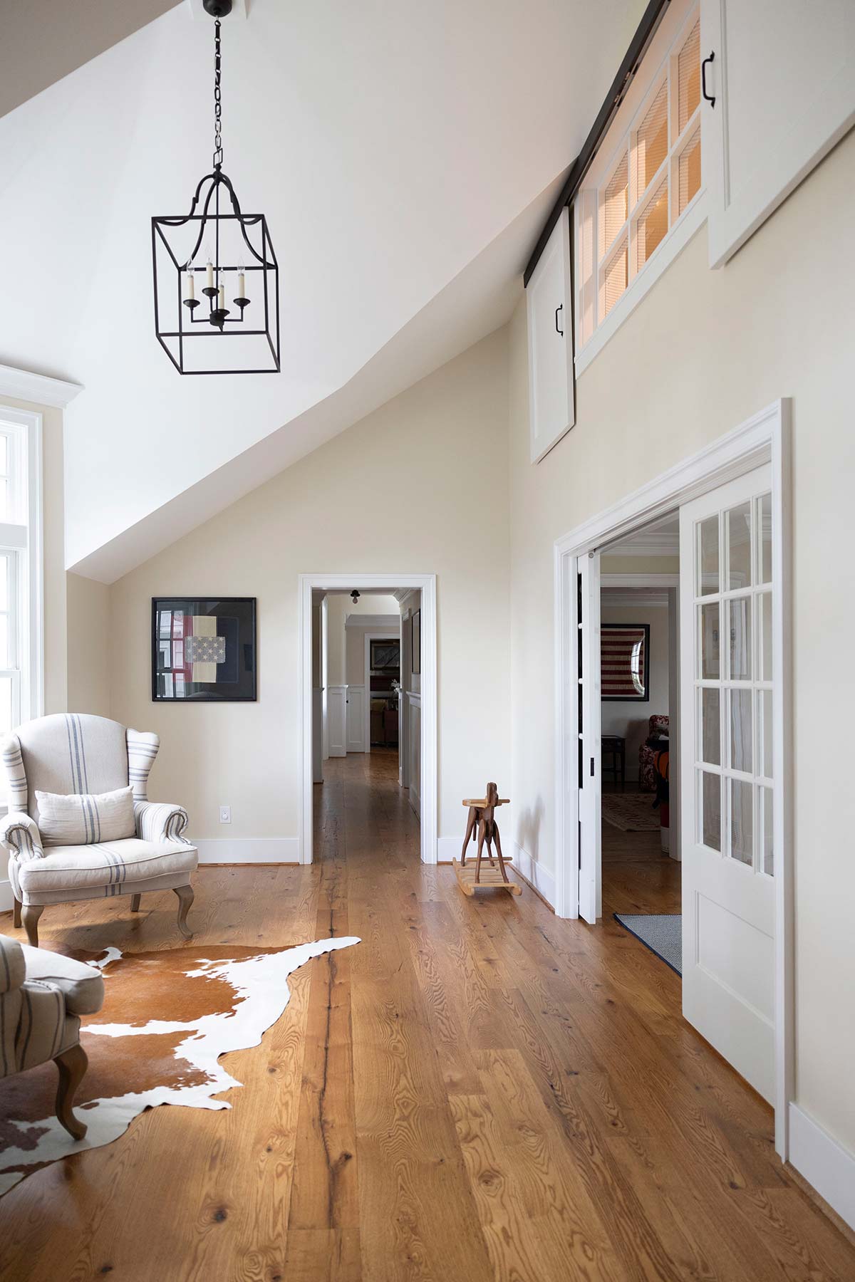 A bright room with sloped ceilings, a chandelier, striped armchair, wooden floors, and glass doors, blending rustic and modern style.
