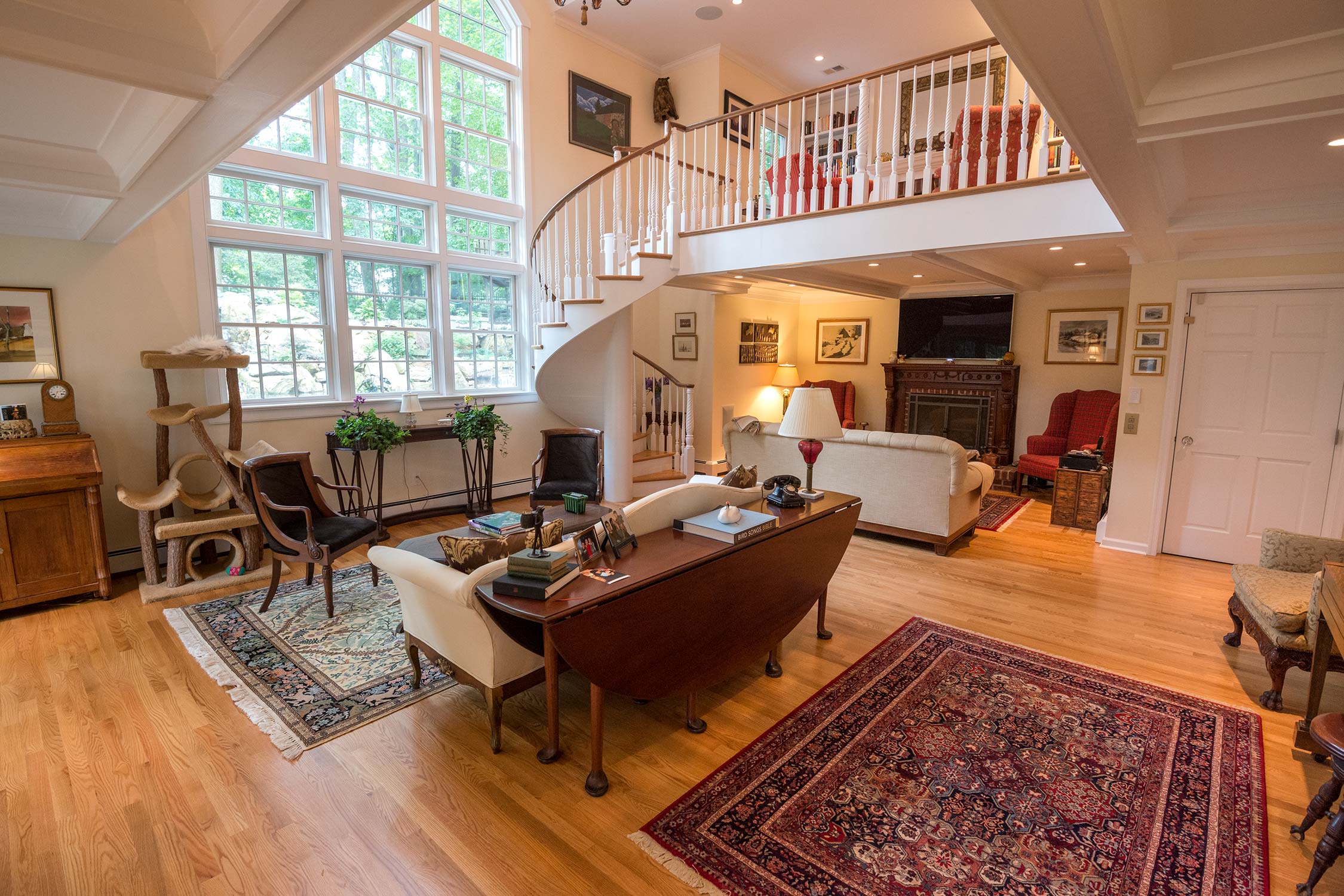A cozy living room with large windows, a spiral staircase, white sofa, red armchairs, wooden table, and a loft area with bookshelves.